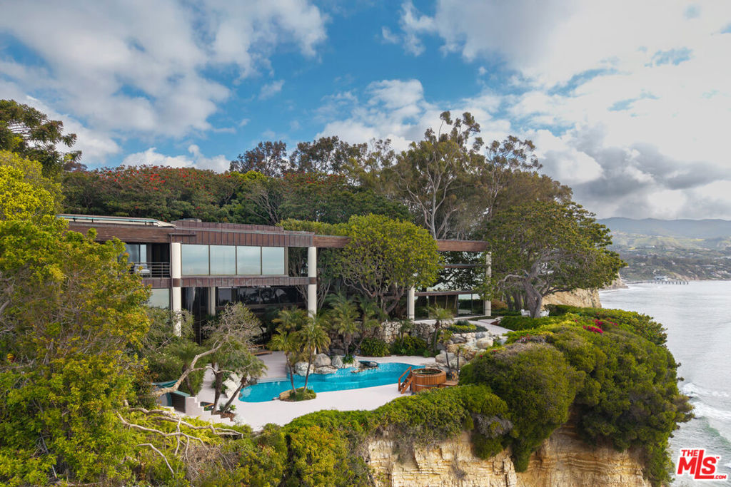 A Point Dume legacy estate of rare provenance and architectural significance, once home to legendary host Johnny Carson, this extraordinary compound spans over four pristine acres across four parcels, commanding panoramic views of the Pacific Ocean, Queen's Necklace, and Paradise Cove. Designed by renowned architect Edward R. Niles, the residence is a sculptural interplay of glass, wood, and geometry, a modernist composition that rises dramatically above the bluffs. At its heart lies an awe-inspiring arboretum pavilion, where a soaring 30-foot glass ceiling latticed with wood slats floods the interiors with natural light. Anchored by a striking copper and glass sunken fireplace encircled by built-in seating, this space seamlessly transitions to bluff-top terraces, a koi pond, and cascading water features. The main level flows effortlessly between dramatic entertaining areas and intimate retreats: a triangular dining room with ocean and pool vistas, a black marble and wood bar lounge, a chef's kitchen with butler's pantry, and a glass-walled office that appears to float among the trees. On the lower level, an ocean-view lounge with wet bar opens to expansive terraces, complemented by a screening room and an impressive wine cellar. Occupying the entire upper floor, the primary suite is a sanctuary of luxury, with dual spa-inspired bathrooms, expansive walk-in closets, two private offices, and a fireplace-adorned bedroom opening to a private terrace with whitewater views.The grounds are an oasis of curated gardens, sweeping lawns, and specimen trees. A saltwater pool with grotto waterfall, Jacuzzi, sauna, and cold plunge invite relaxation, while al fresco dining terraces capture the drama of Malibu sunsets. Additional estate features include a circular glass security office with kitchenette and bath, a staff cottage, and motor facilities with three-car garage, Tesla chargers, and capacity for up to 40 vehicles. Across a separate parcel lies a championship tennis complex, framed by manicured rose gardens and sculpture installations. The travertine tennis pavilion offers two guest suites, a full kitchen, fireplace lounge, spa-style locker rooms with sauna, and expansive courtside terraces. Additional guest accommodations include a private guesthouse, greenhouse, fruit orchard, and aviary.A true Malibu icon, 6962 Wildlife Road embodies the convergence of architectural artistry, land, and legacy, creating an estate of unrivaled beauty, privacy, and prestige.