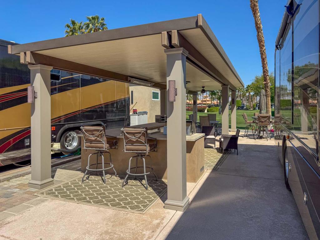 This luxury RV lot at Outdoor Resort Indio boasts stunning golf course views, an extended alumawood pergola with a solid roof, and a spacious outdoor kitchen, creating the perfect setting for upscale resort living. The large L-shaped kitchen, complete with stainless appliances and a built-in bar, sits under the pergola, offering both shade and comfort. Two ceiling fans and two ceiling heaters ensure year-round enjoyment, while the paver lot extensions provide extra space for relaxation and entertainment. An eight-chair dining table and a four-seat tall table with an umbrella offer ample seating, making this lot ideal for hosting and unwinding in style.Outdoor Resort Indio is a Class A only RV resort in Indio, CA.