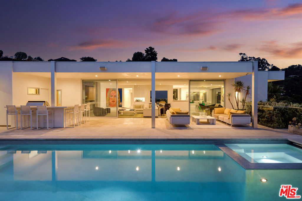 Set behind a sleek white brick facade and elevated above the street, this meticulously updated mid-century residence commands attention in the heart of Trousdale Estates. A series of floating stone steps, framed by sculptural desert landscaping and uplighting, leads to a private gated entry that opens into a serene front courtyard. The architectural silhouette is clean and offers a strong sense of privacy while introducing a refined, modern edge. From the moment of entry, glass walls offer glimpses through to the panoramic vistas beyond, setting the tone for a home defined by light, openness, and connection to its surroundings. Inside, an expansive open layout unfolds across multiple living and entertaining areas, each thoughtfully positioned to emphasize flow and connection to the outdoors. The formal living room, anchored by a clean-lined fireplace, opens seamlessly to the patio and grounds beyond. Adjacent is a separate dining room designed for intimate dinners or larger gatherings, complemented by a chef's kitchen outfitted with top-tier appliances and a breakfast area that overlooks the hills. The primary suite is a sanctuary of calm, with sparkling city light views, direct access to the outdoors, and an ensuite bathroom featuring a soaking tub and glass-wrapped shower set against an extraordinary skyline backdrop. An additional guest suite offers its own bathroom and private outlook, while a third bedroom, ideal for staff, includes an ensuite bath. A flexible den or office features its own bathroom and serves as a convenient pool bath as well. The outdoor living experience is equally considered, with limestone floors carried from inside to out, creating continuity between environments. A built-in barbecue and kitchen are flanked by bar seating for six, while a covered lounge and dining area extend the living space under open skies. Every element of the home inside and out reflects its mid-century roots while embracing contemporary updates designed for comfort, elegance, and effortless indoor-outdoor living. Heightened by its position in the iconic Trousdale Estates, sweeping, unobstructed views from downtown to the Pacific serve as a constant reminder of its place at the pinnacle of Beverly Hills living.