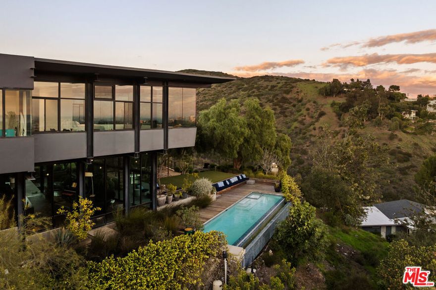 Designed and built as the personal residence of Hagy Belzberg, FAIA, this exceptional 5,000 square foot architectural home is a rare expression of a master architect building for himself. Belzberg's work, recognized internationally through decades of award-winning residential, cultural, and hospitality projects, has long explored the intersection of experimentation and livability. That philosophy is fully realized here, in a home conceived and built for personal use, free from the constraints of speculation, where every decision reflects the priorities of an architect designing for his own life. Set privately and securely, in a gated 4-home enclave, on a 2- acre lot with sweeping city and canyon views, the residence unfolds as a sequence of carefully calibrated spaces defined by steel framing, warm wood surfaces, and expansive walls of glass. Light, proportion, and circulation are methodically considered, creating a home that is both aesthetically singular and deeply comfortable. The main living level is conceived for both everyday living and sophisticated entertaining. Floor-to-ceiling walls of glass  dissolve the  boundaries between interior and exterior, opening to terraces, gardens, and a dramatic infinity-edge pool that appears to float above the landscape. Living and dining spaces flow effortlessly into a design-forward kitchen with custom millwork, professional-grade appliances, and generous storage integrated seamlessly into the architecture. Throughout the home, bespoke detailing reflects the sensibility of an architect's own residence: integrated shelving and libraries, curated sightlines, and thoughtful transitions between public and private zones. A private media room offers a cinematic retreat, while a dedicated gym provides a discreet wellness space, and a 3-station library/study area invites educational and occupational advancement. The primary suite is a serene sanctuary, framed by panoramic views and anchored by warm materials and clean lines. A spa-like bath, generous closets, and a sense of calm separation complete the experience. The three additional bedroom suites maintain the same level of design achievement and comfort. The outdoor spaces are equally intentional: layered terraces, intimate garden moments, and seating areas designed for both gathering and solitude. As day turns to dusk, the home glows from within, the architecture revealing its structure and rhythm against the evening sky. More than a residence, this is a self-authored work of architecture; a home conceived without compromise, where innovation serves livability, and design is meant to be inhabited, not merely admired. Located moments away from the coveted dining, nightlife and shopping on San Vicente and in Brentwood Country Mart and within some of the city's most highly acclaimed public schools (Kenter Canyon Elementary Charter, Paul Revere Charter Middle School and Palisades Charter High School).