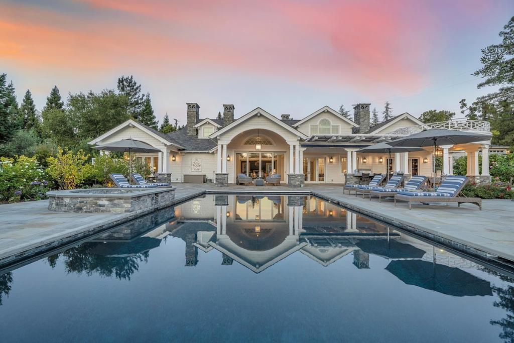 At the end of a private lane, this gated Hamptons-inspired residence pairs timeless architecture with modern sophistication. Over 6,000 sf of living space showcases generous rooms, high ceilings, crisp white custom millwork, oak floors, and designer lighting throughout. A welcoming formal entry opens to elegant living spaces including a gourmet kitchen with Italian Bardiglio marble island, Sub-Zero and Wolf appliances, dining area, and large family room with retractable glass doors to an expansive terrace. The formal living room offers a tray ceiling, stately fireplace, and patio access, while a dramatic two-story library with loft and alder cabinetry provides a flexible retreat. The 5-bedroom en suite layout includes 3 suites on the main level, a spectacular primary with spa-like bath, and 2 upstairs suites with an additional family room and private entry. Resort-style 1.1-acre grounds feature an infinity pool, spa, outdoor kitchen, fire pit, gardens, vineyard, wine cellar, and saunaan extraordinary setting for everyday living or entertaining.