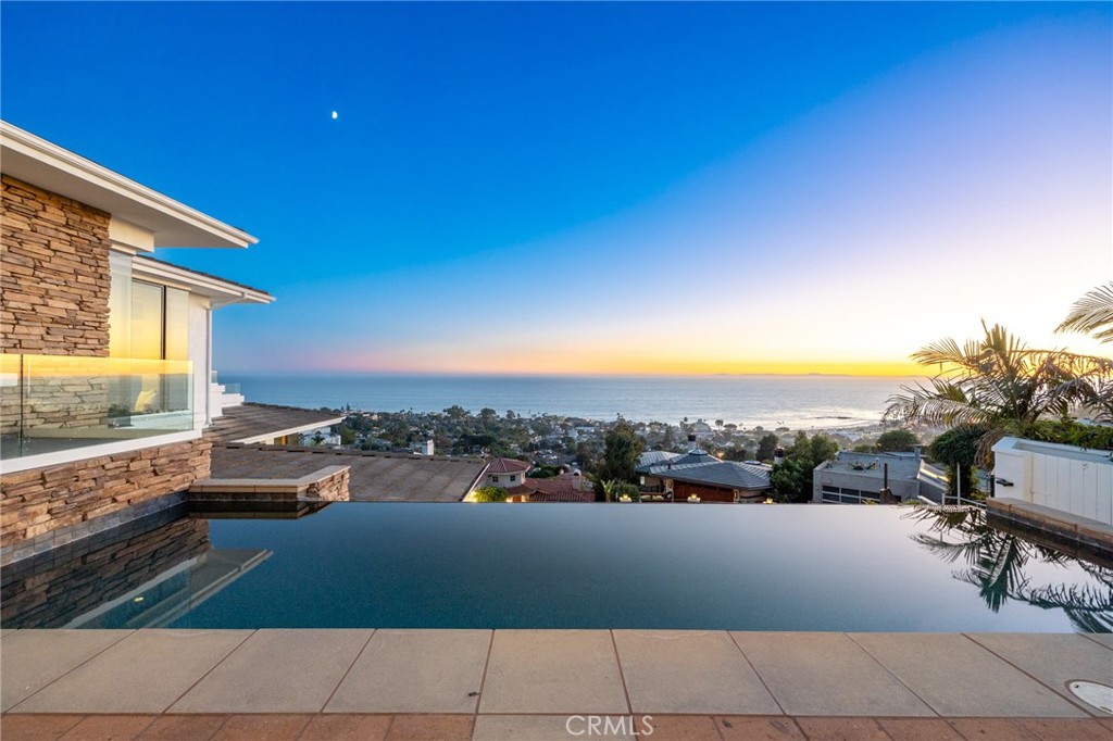 Perched in one of the world’s most celebrated coastal destinations, this rare estate-sized residence in Laguna Beach offers a lifestyle of unmatched luxury and breathtaking beauty. Just blocks from the beach and the vibrant Village, the home commands sweeping panoramic views of Catalina Island and iconic Main Beach—an extraordinary vantage point few properties can claim. Spanning over 6,000 square feet of refined living space, this gated estate rests atop a generously elevated one-third acre lot, designed to maximize privacy and showcase the stunning seascape. The exterior features an infinity-edge pool that seemingly spills into the Pacific, a three-car attached garage, and a multi-vehicle motor court, all framed by sculpted hardscape and a curated palette of coastal succulents. Inside, the home is a masterclass in architectural sophistication and custom craftsmanship. Highlights include: a gourmet chef’s kitchen with premium finishes, a glass elevator offering ocean views on every ride, rich teak flooring throughout, a dedicated home theater for cinematic indulgence, and fireplaces in the living room, primary suite, and outdoor terrace—perfect for cozy evenings by the pool. The layout features three luxurious bedroom suites and a spacious office that easily converts to a fourth bedroom, offering flexibility for guests or remote work. Smart home amenities abound, including: integrated stereo speakers throughout, a sophisticated lighting control system, dual-zone heating and air conditioning, and comprehensive security and central vacuum systems. Powered by state-of-the-art solar panels, the residence delivers energy-efficient living while dramatically reducing utility costs. From the first light of dawn to the golden hues of sunset, this home captures the essence of Laguna Beach in every moment. Indulge in coastal elegance—where every detail is designed to elevate your lifestyle.