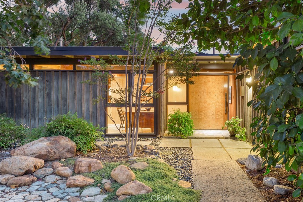 This extraordinary residence is a Mid-Century Modern post-and-beam masterpiece, designed by Everett Tozier & reimagined by Hartman Baldwin with uncompromising attention to detail. A sprawling front yard enveloped by lush, curated landscaping creates a private sanctuary & sets the tone for the home’s refined elegance. The striking custom pivot door made of walnut & ash-olive wood offers a grand welcome, revealing interiors where dark slate tile flows into warm white oak floors, a pairing that harmonizes beautifully with the home’s architecture. A dramatic floor-to-ceiling wall of glass floods the open living & dining rooms with natural light, framing verdant views while highlighting the granite fireplace’s bold textures. The dining room flows effortlessly into a showpiece kitchen designed for both culinary artistry & social connection. Crafted with custom birch cabinetry, the space pairs sleek stone countertops with a sophisticated backsplash for visual depth. High end appliances, including a built-in refrigerator, offer both beauty & function, while the layout invites effortless entertaining with open sightlines to the living & dining rooms. Exposed beams & fine finishes stay true to its post-and-beam heritage, creating a space that feels both timeless & modern. Three spacious bedrooms & two exquisite bathrooms include a primary suite transformed into a true retreat, complete with a sitting room featuring built-in bookcases, a pocket door for privacy & serene backyard views. This en suite is a luxurious escape with a granite slab shower, custom vanity & expansive walk-in closet. Dual outdoor access connects the primary to both the backyard & the poolside patio. While the main house offers 2,154 SF, the guest house at 420 SF, seamlessly integrates with the carport, offering its own open layout with a kitchenette, living room, bedroom & bathroom, perfect for extended stays or private hosting. Expansive & beautifully designed, the backyard offers multiple lounging & dining areas framed by mature trees & manicured greenery. From shaded retreats to open sunlit spaces, it provides the perfect setting for both serene relaxation & lively gatherings. A shimmering pool sits beside the primary suite, creating a resort-like ambiance & a stunning focal point for the outdoor living experience. A property of rare architectural significance & thoughtful craftsmanship, this home delivers an unparalleled lifestyle where design, comfort & privacy converge in perfect harmony.