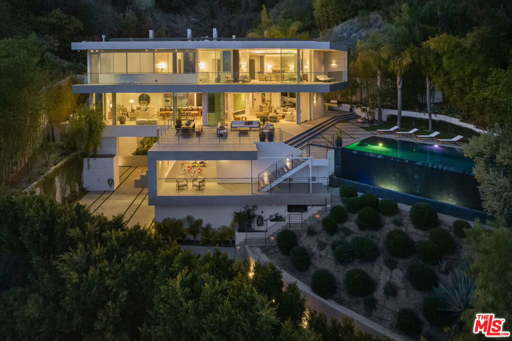 Nestled in the prestigious Hollywood Hills on famed Rising Glen, this stunning contemporary estate offers the perfect blend of modern luxury and natural beauty. Designed with seamless indoor-outdoor living in mind, the residence features expansive floor-to-ceiling glass doors that frame breathtaking panoramic city views. The open-concept living space is anchored by a sleek, state-of-the-art kitchen with custom cabinetry, high-end appliances, and a spacious island that flows effortlessly into the dining and lounge areas. Elegant finishes, warm wood tones, and sophisticated marble accents add to the home's refined ambiance. The primary suite is a private sanctuary, complete with a fireplace, direct balcony access for uninterrupted sunset views, and dual spa-inspired bathrooms - one featuring a soaking tub and the other a spacious glass-enclosed shower along with separate walk-in closets for ultimate comfort and privacy. Additional bedrooms are designed for both style and relaxation, while a dedicated office, home gym, and lounge spaces provide the perfect setting for productivity and leisure. Step outside to a resort-inspired backyard featuring an infinity-edge pool, a serene spa, and multiple lounge areas designed for entertaining or unwinding in privacy. Lush landscaping and towering palms enhance the sense of tranquility, while an intimate outdoor courtyard offers a secluded retreat. With a flawless blend of architectural brilliance and contemporary elegance, this home is a true masterpiece, offering an unparalleled living experience in one of L.A.'s most sought-after neighborhoods.