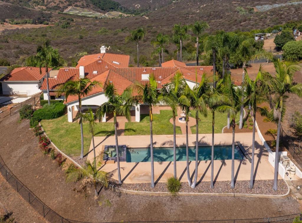 Set atop a hill on 3.06 acres in the heart of horse country, this Mediterranean estate in coveted Hialeah Estates enjoys sweeping 360-degree views of rolling countryside and coastal breezes. Just 2.7 miles from the San Luis Rey Training Center, the property was designed to celebrate its extraordinary setting, with expansive windows and effortless indoor-outdoor flow to a backyard retreat complete with pool, spa, and lush landscaping framed by palm trees. The inviting floor plan offers formal living and dining rooms, three bedrooms on the main level and a secluded primary suite in its own upper wing, while the open-concept kitchen, breakfast area, and family room with fireplace create a warm hub for both relaxed living and stylish entertaining. Additional amenities include a refrigerated wine room, an attached three-car garage, and a detached two-car garage—delivering a perfect balance of elegance, comfort, and functionality in a truly breathtaking location.