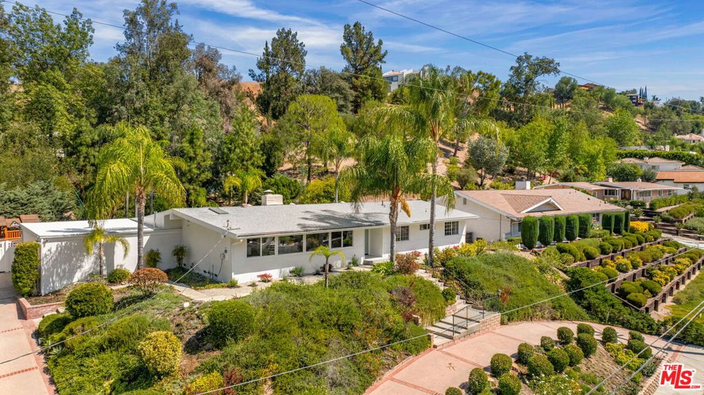 Perched gracefully above the street in one of Woodland Hills' most desirable South of the Boulevard enclaves, this beautifully reimagined residence offers total privacy and resort-style living. Step inside to discover a light-filled great room with vaulted ceilings, white oak engineered hardwood floors, and walls of glass that open to views of the sparkling pool. A double-sided fireplace anchors the space, seamlessly connecting the living and dining areas with the open chef's kitchen, an entertainer's dream. The backyard is a true oasis: a sun-drenched pool and spa, a large flat grassy lawn with play structure. A wooded area at the back of the property ensures unmatched privacy and an incredible backdrop to your outdoor living. Additional highlights include a converted garage that functions as a private office, playroom, or flex space or which could easily be converted back to a garage. The exterior of the home is beautifully finished flat Santa Barbara stucco. Perfectly positioned near acclaimed Calabash Charter Elementary and moments from the shops, dining, and canyon trails of Woodland Hills.