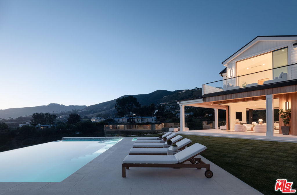 Welcome to this ultra-private, 2025-built Modern Farmhouse, gated and positioned far from the street beyond a long meandering driveway in one of the best sections of Malibu. Complete with a full freestanding guest house/ADU and boundless park-like grounds meticulously designed by renowned landscape architects, the home offers sweeping views of the ocean, hills, and Trancas Canyon, all set above Zuma Beach and Malibu High School. Discover high-quality finishes throughout this open floor plan, optimized for an effortless indoor-outdoor lifestyle. Soft textures of Italian limestone and smooth wood panels complement rich dark steel grays and traditional coastal siding. A serene double-height foyer is open and airy, with huge panes of glass welcoming warm natural light within as wide-plank oak floors flow into the public entertaining spaces. The gourmand's kitchen, central to the open-concept layout, is ideal for catering and serving party guests, fitted with chef-grade appliances including a Wolf range and Miele espresso station, large center island with prep sink, Cararra marble counters and backsplash, and tasteful reeded accents. Nearby, the living room is highlighted by a Brutalist travertine fireplace flanked by custom built-ins that are as beautiful as they are functional. Corner-free pocket Styline doors retreat for optimal transition to the grassy backyard, including an infinity pool, spa, cannonball fire table, outdoor barbecue, dining, and mesmerizing vistas of the Santa Monica Mountains and vibrant blue Pacific. Upstairs, the lavish primary suite includes an owner's living room, dual walk-in changing rooms, and an ocean-facing wraparound balcony connecting the bedroom to the spa-inspired bathroom, complete with soaking tub, makeup desk, and double-size rainwater shower encased in bookmatched stone. Downstairs is a quiet lounge, perfect for use as a media room, gaming room, or billiards room which opens to the outside. The guest house has a full kitchen, dining and living, and one bedroom plus bathroom. There is also an outdoor-facing bathroom with shower and a finished garage that includes a mud room area. Thoughtfully curated drought-tolerant landscaping is peppered throughout the 1.48-acre usable flat grounds, its blooms attracting colorful butterflies and other natural pollinators. Nearby homesteads along the hills offer a delightful showing of various happy farm animals such as pigs, goats, horses, and mules. This is scenic coastal living at its very finest, just moments to popular surf hot spots such as Leo Carrillo and County Line as well as Point Dume, Pepperdine, and the Malibu Colony and Country Mart.
