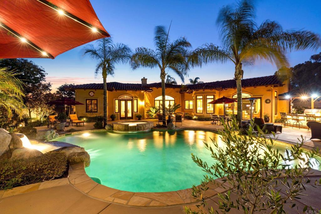Welcome to 12105 View Pointe Row, an elegant single-level estate in the private 5-home gated community of View Pointe Row in Rancho Bernardo. Built in 2002 and meticulously maintained, this 4,434 sq ft, 4-bedroom, 3.5-bath home blends timeless Spanish Mediterranean design with modern energy efficiency, including owned solar for both the home and pool plus two Tesla batteries. Set on a picturesque lot overlooking the 4th hole of The Heights Golf Club, this residence offers sweeping mountain and fairway views. Inside, the open floor plan is enhanced by arched ceilings, gorgeous crown molding, travertine and hardwood flooring, and additional custom finishes throughout. The gourmet kitchen features granite counters, a walk-in pantry, and top-of-the-line appliances, flowing seamlessly into the spacious family room with its dramatic stone fireplace, entertainment center, and wet bar with wine storage. The primary suite is a private retreat, complete with its own fireplace, a spa-inspired Jacuzzi bath with dual vanities, and a massive walk-in closet. Resort-style living continues outdoors in a backyard designed for entertaining and relaxation. A pebble-tech pool and spa with waterfall is complemented by a covered cooking area and bar top, and generous patio space. Lush landscaping with accent lighting, a rose and herb garden, turf lawn, and drip irrigation system complete the grounds. Additional highlights include a 4-car garage with epoxy flooring, dual-zoned heating and A/C, and Google-integrated speakers and smart-home controls throughout. This pristine home offers a rare opportunity to enjoy single-level luxury living with panoramic views in one of Rancho Bernardo’s most desirable gated enclaves.
