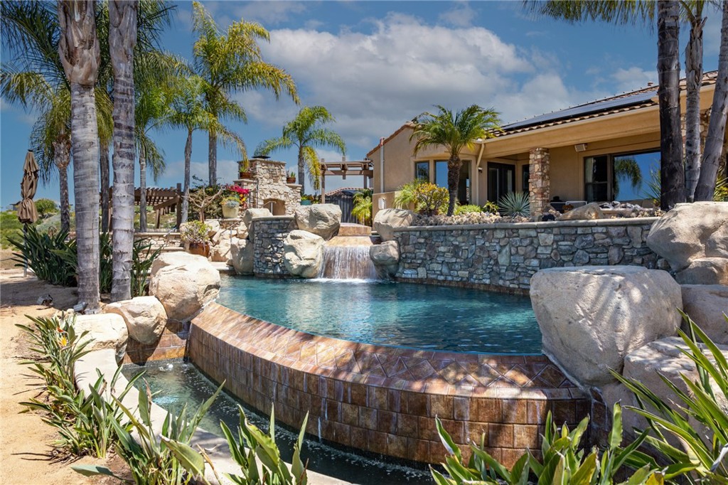 Reduced!! Owner will carry First Deed of Trust! Do not miss this gorgeous home nestled in Fallbrook's highly desirable Sycamore Ranch community, where the Golf Club of California is just a swing away! Perched on 2.34 glorious acres, this single-story dazzler sprawls over 4,600 square feet and serves up jaw-dropping views of San Luis Rey Valley, neighboring vineyards, and the majestic west mountains. The facade boasts a 3-car garage with snazzy epoxy floors, an EV charger, and a curvy driveway fit for your RV or boat. On the south side of the lot, discover a 3,000 sqft wonderland perfect for epic events to set the stage for alfresco fun with views that'll knock your socks off! There are 2 lots with separate addresses ideal for an ADU or extra Casita, plus easy access to neighboring horse trail.
In the backyard, dive into a solar-heated pebble tec saltwater pool, unwind in the gas-heated spa, or cozy up by the gas fireplace and fire ring. Entertainment zones abound with tiered lounges and a covered patio. With several avocado trees,2.34 acres of outdoor space and endless possibilities, your family's outdoor dreams await! Enter through grand double doors into a private courtyard adorned with pavers, a wood-burning fireplace, and stone veneered columns. The Casita (5th bedroom) boasts its own entrance, bath, climate control (A/C and heat), and hardwood floors. Inside, 12' ceilings and hardwood floors lead you to a formal dining room with vineyard vistas and a family room featuring a custom stone veneer fireplace and surround sound.
The kitchen wows with maple cabinetry, a one-level island with a prep sink, breakfast bar, walk-in pantry, Butler's pantry and custom glass-blown drop lights. Feast your eyes on GE's built-in microwave and a KitchenAid appliance suite: 48" gas stove with griddle, dishwasher, double ovens, and a double-door built-in 48" fridge. The primary suite is a sanctuary, complete with space for a Cal King bed, a cozy seating area, a fireplace and sweeping views. The en-suite bath pampers with double vanities, walk-in closets, a large shower, tub and gorgeous privacy barn door. All bedrooms are nice sized with en-suite bathrooms. Big ticket items are all set: 3 a/c units, a 200 amp panel, owned 40-panel solar system (14.4 kilowatts), a solar pool heater, a tankless water heater (with recirculating pump), and new LED lighting throughout. Solar lighting tubes light up the home with zero energy use.