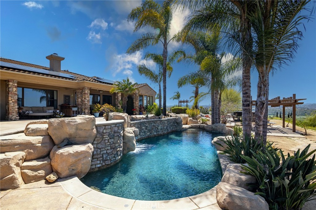 Reduced!! Owner will carry First Deed of Trust! Do not miss this gorgeous home nestled in Fallbrook's highly desirable Sycamore Ranch community, where the Golf Club of California is just a swing away! Perched on 2.34 glorious acres, this single-story dazzler sprawls over 4,600 square feet and serves up jaw-dropping views of San Luis Rey Valley, neighboring vineyards, and the majestic west mountains. The facade boasts a 3-car garage with snazzy epoxy floors, an EV charger, and a curvy driveway fit for your RV or boat. On the south side of the lot, discover a 3,000 sqft wonderland perfect for epic events to set the stage for alfresco fun with views that'll knock your socks off! There are 2 lots with separate addresses ideal for an ADU or extra Casita, plus easy access to neighboring horse trail.
In the backyard, dive into a solar-heated pebble tec saltwater pool, unwind in the gas-heated spa, or cozy up by the gas fireplace and fire ring. Entertainment zones abound with tiered lounges and a covered patio. With several avocado trees,2.34 acres of outdoor space and endless possibilities, your family's outdoor dreams await! Enter through grand double doors into a private courtyard adorned with pavers, a wood-burning fireplace, and stone veneered columns. The Casita (5th bedroom) boasts its own entrance, bath, climate control (A/C and heat), and hardwood floors. Inside, 12' ceilings and hardwood floors lead you to a formal dining room with vineyard vistas and a family room featuring a custom stone veneer fireplace and surround sound.
The kitchen wows with maple cabinetry, a one-level island with a prep sink, breakfast bar, walk-in pantry, Butler's pantry and custom glass-blown drop lights. Feast your eyes on GE's built-in microwave and a KitchenAid appliance suite: 48" gas stove with griddle, dishwasher, double ovens, and a double-door built-in 48" fridge. The primary suite is a sanctuary, complete with space for a Cal King bed, a cozy seating area, a fireplace and sweeping views. The en-suite bath pampers with double vanities, walk-in closets, a large shower, tub and gorgeous privacy barn door. All bedrooms are nice sized with en-suite bathrooms. Big ticket items are all set: 3 a/c units, a 200 amp panel, owned 40-panel solar system (14.4 kilowatts), a solar pool heater, a tankless water heater (with recirculating pump), and new LED lighting throughout. Solar lighting tubes light up the home with zero energy use.