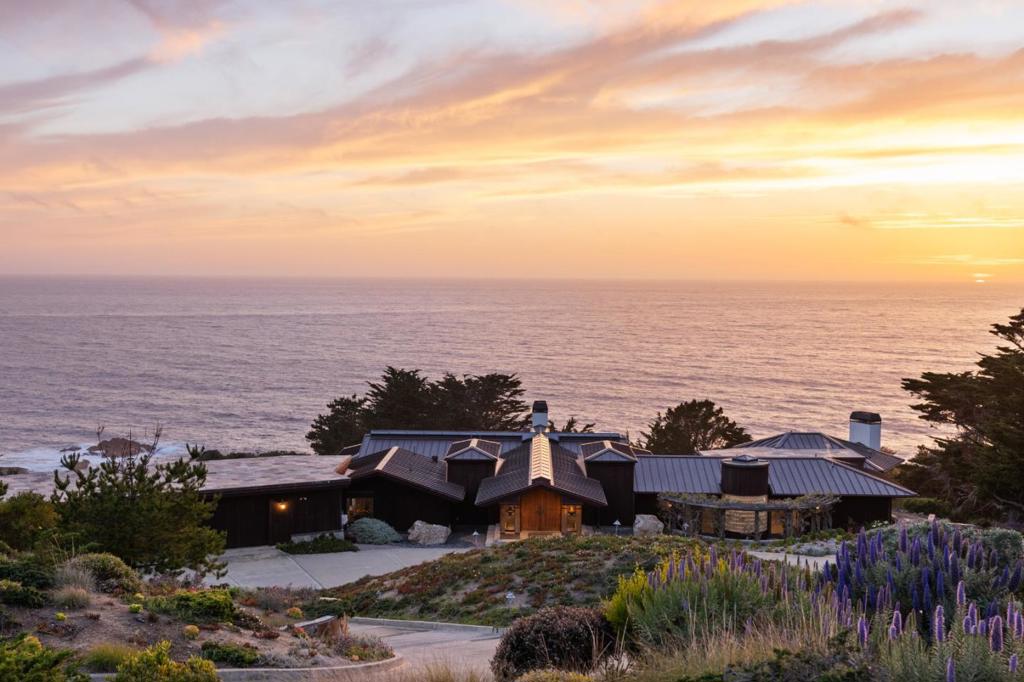 Modern elegance framed by coastal serenity. This exquisite four-bedroom, six-bathroom estate is nestled between the Santa Lucia Mountains and the Pacific Ocean, offering nearly six acres of privacy and sweeping views of the Carmel coastline. Sunlight pours through skylights and vast windows, illuminating a vaulted living room that flows into an elegant dining space. The gourmet kitchen features top-tier appliances, custom cabinetry, a sleek breakfast bar, and a cozy dining nook ideal for morning rituals. Step onto the wraparound balcony and breathe in the salt-kissed breeze as the ocean unfolds before you. The primary suite offers a gas fireplace, wood-clad ceiling, and a spa-like bath with cedar sauna and luminous glass block shower. A generous home office lies just beyond. Descend the spiral staircase to a recreation lounge with state-of-the-art theater, wine cellar, and wet bar. Two guest suites, each with private bath and walk-in closet, extend a warm welcome. A three-car garage, dual laundry rooms, and a private-access studio with kitchenette and bath complete this timeless sanctuary by the sea.
