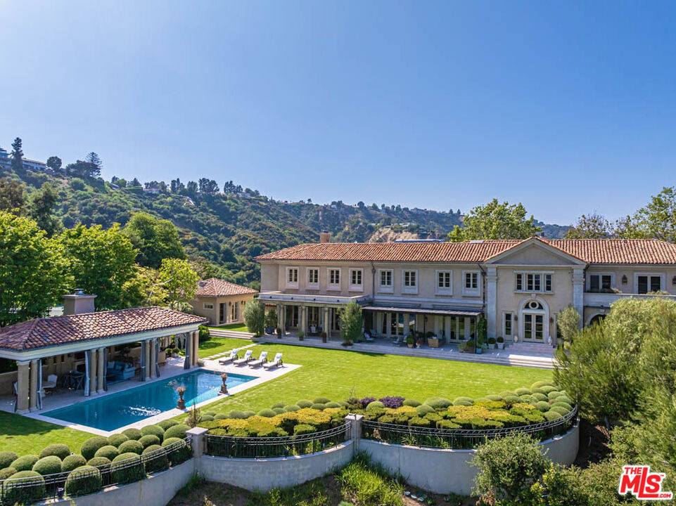 Enchanting Bel-Air Villa: A Spectacular Retreat Above the City. Nestled behind discreet gates, perched above the bustling cityscape, lies the enchanting Bel-Air Villa, offering breathtaking panoramic vistas. A private, meandering road, flanked by towering, verdant hedges, leads to the grand motor court. Step into the realm of architectural mastery crafted by Oscar Shamamian, where the facade adorned with romantic, climbing vines beckons. Upon entry through the voluminous 2-story foyer, be captivated by the picturesque city vistas that unfold. Renowned designer Michael Smith has curated the interiors to perfection. Limestone flooring guides one to a stately formal living room boasting lofty ceilings, a majestic stone fireplace, and French doors opening onto a charming veranda. Adjacent, a sun-kissed office adorned with bespoke shelving and a terrace awaits. The generous formal dining room, with its white wood-beamed ceiling and intricate crown molding, exudes elegance with textured wallpaper accents. Ascend the architecturally unique stacked staircase to discover the sumptuous primary bedroom suite, featuring a fireplace, wrap-around terrace, and not one, but two luxurious bathroom suites. Her sanctuary boasts a glamorously appointed soaking tub, a fireplace, and vaulted exposed wood beam ceilings, offering tranquil views. His retreat features a marble bathroom, patio access, and a spacious walk-in closet. Completing the upper level are four additional bedroom suites, accessed via a discreet back staircase leading to a striking gallery bathed in dramatic lighting and glass doors opening onto a terrace with serene canyon vistas. The heart of the home, the gourmet chef's kitchen, delights with a central island, top-of-the-line appliances, a butler's pantry, and a quaint breakfast nook. Adjacent, a spacious family room beckons with a cozy fireplace, built-in bar, and a discreetly concealed projector screen. Outside, the vibrant grounds invite alfresco dining and lounging amidst a profusion of blooming flowers, verdant foliage, and bountiful herbs, evoking the charm of an English country garden. Entertainment abounds with a swimming pool, pool pavilion boasting a fireplace and built-in barbeque, and a spa offering sweeping views. Luxurious amenities abound, including a 3-car garage, staff quarters, a guest house, and a championship North-South tennis court. This magical retreat offers a spectacular retreat within the heart of Bel-Air.