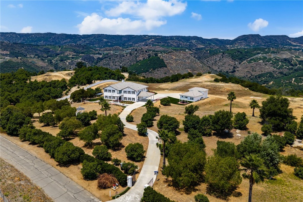 Welcome to your own private hilltop retreat—an awe-inspiring estate set on over 25 acres of mostly flat land, offering unparalleled vistas that will astound you. A grand entrance through stately wrought iron gates leads you along a rose-lined driveway to this spectacular residence. Surrounded by a thriving avocado grove, this property provides a peaceful, picturesque setting perfect for those seeking elegance, privacy, and a connection to nature. Chef’s Dream Kitchen equipped with premium Viking appliances, walk-in pantry with custom stained-glass door, and elegant quartzite countertops, 4 Spacious Bedrooms—each designed as a luxurious retreat, 4 Impeccably Designed Bathrooms featuring high-end finishes and meticulous attention to detail. Upgraded Flooring Throughout and two cozy fireplaces. Central Heating and Air for year-round comfort. Enjoy two expansive balconies on the upper level, offering a front-row seat to stunning sunrises, sunsets, and rolling fog over the hills—an ideal space for entertaining or quiet morning coffee. Experience true tranquility and inspiration from this hilltop haven. Perfect for equestrian enthusiasts, nature lovers, farmers, or anyone in search of a rare lifestyle opportunity. The existing avocado grove provides both serenity and potential income, with room to expand for even greater productivity. Located just 15 minutes from the charm of Old Town Temecula, this one-of-a-kind estate seamlessly blends luxury living with natural beauty.