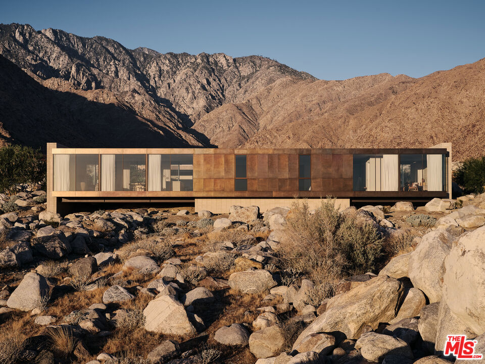The Woods House, 2021, Woods + Dangaran. From siting to floor plan to finishes, this residence is deeply connected to the desert environment and its context in the hills above Palm Springs. Surrounded by boulders and spanning a natural arroyo, the home is lifted above existing site features, yet still retains a powerful connection to the earth. The architecture emphasizing horizontal lines, natural materials, and visual and physical linkages between indoors and out celebrates and elevates the tenets of desert modernism. Exterior walls of earth-toned plaster and textured concrete masonry units tie the slab-on-grade structure to the site. Deep roof overhangs are wrapped in brass paneling. Strategically-placed floor-to-ceiling glazing frames stunning views of the desert in all directions. A glass-enclosed bridge connects two wings of the home and creates a central atrium that can be viewed from nearly every room. Large sliding glass doors run the length of the great room and provide an uninterrupted transition to an outdoor dining area, deck, pool, spa, fireplace, and lounge. Lot 2 APN 504-390-002 to be included in sale. Renderings for the second parcel by Woods + Dangaran available by request.