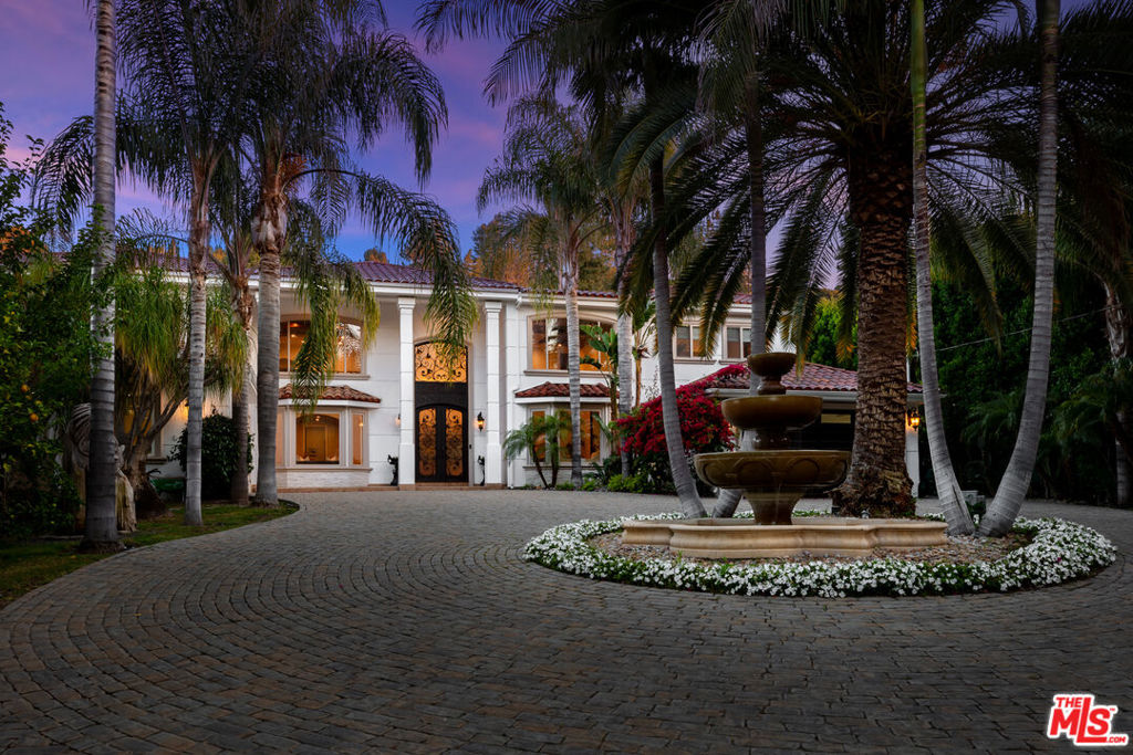 Secluded behind privacy gates amidst towering palms and drought-tolerant landscaping is this regal Mediterranean estate on more than half an acre of lush park-like grounds, positioned on a quiet part of Valley Vista south of Ventura Boulevard. An expansive cobblestone motor court is centered by a tranquil European fountain, affording an abundance of parking for guests. Vibrant bougainvillea complements the exterior's ornamental details as an ornate wrought-iron door welcomes visitors upon arrival. Inside, a chandelier glistens above the double-height foyer as decorative railing and a shimmering charcoal accent wall serve as backdrop. The formal living room is complete with a chateau-worthy fireplace and bay window near the formal dining room, with its coffered ceilings high above and large picture window inviting sunlight within. Tile flooring continues into the family room, appointed with a built-in media center, exposed cross-hatch beams, wet bar, and floor-to-ceiling panes of glass that open to the outside. The kitchen is dressed in warm woods and offers a delightful iridescent backsplash, copious counter prep space, eat-in breakfast area, and walk-in pantry. State-of-the-art appliances include a Thermador cooktop and warming ovens, Sub-Zero refrigerator, and commercial-grade freezer. Upstairs, discover a lavish primary suite with large sitting area, two-sited fireplace, and arched French doors that open to a lengthy wraparound balcony overlooking the paradisal rear grounds. The primary bathroom has an oversized jetted tub, glass-enclosed shower, built-ins, and dual vanities, one of which features a makeup desk. Secondary bedrooms have generous closets, and bathrooms have custom finishes. Enjoy a fresh all-needs-met lifestyle from numerous living spaces including a billiards/card room, 12-seat theatre, office/library, and gym. Outside, relish a totally private resort-like sanctuary with pool, spa, outdoor kitchen and barbecue, fire pit, and multiple patios for lounging, dining al-fresco, and relaxing under California skies. A meandering stone pathway leads to a gazebo surrounded by verdant plantings, grassy lawns, and blossoming flora, ideal for casual conversation on warm summer nights. A sports court offers a variety of recreational and competitive possibilities including tennis, pickleball, basketball, and more. Welcome to this prime, coveted section of Encino with close proximity to award-winning Lanai Road Elementary, parks, golf courses, and major freeways providing Westside/DTLA access, just moments to the many shops, boutiques, and eateries along Ventura Boulevard.