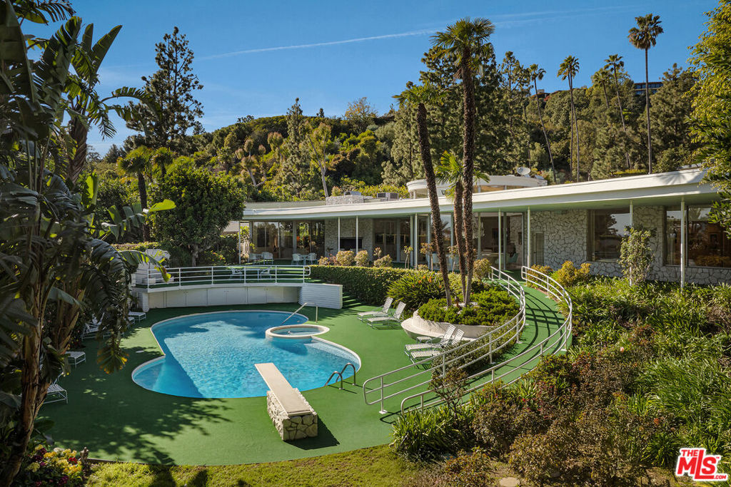 Hitting the market for the first time since its creation in 1959, this exceptionally rare estate in Trousdale Estates is a true generational offering. Set on over an acre in one of the most coveted enclaves of Beverly Hills, this property presents a once-in-a-lifetime opportunity to own a residence of remarkable provenance and scale. Originally built by the sellers, the estate occupies an unrivaled position within Trousdale, offering unparalleled privacy, city views, and the chance to reimagine a mid-century masterpiece in one of the most sought-after locations in Los Angeles. A grand circular entryway, bathed in natural light from a soaring skylight and adorned with a dazzling crystal chandelier, transports you to the golden age of mid-century elegance. The moment you step inside, time slows, and the home's original glamour unfolds a masterpiece of an era where design was bold, details were deliberate, and every arrival felt like an occasion. The front living room offers a warm and inviting ambiance, complete with a fireplace and sliding doors that open onto an expansive outdoor patio deck. Beyond, the entry unfolds into a magnificent entertaining space centered around a circular sunken bar, positioned beneath a breathtaking oversized crystal chandelier. Encircled by skylights, this architectural centerpiece allows sunlight to dance throughout the day, casting elegant cascading shadows and filling the space with an ever-changing glow. The expansive formal living room features multiple sitting areas, seamlessly blending indoor and outdoor living through floor-to-ceiling glass doors.The formal dining room is illuminated by an exquisite crystal chandelier, with floor-to-ceiling windows framing serene views of a lush flower garden and a striking gallery-style waterfall fountain installation. A chef's kitchen designed for both communal gatherings and functionality, features a family room, office area, separate staff quarters, dedicated prep area, and dual laundry room - all with access to the backyard lawn and vegetable garden. The primary suite offers a private sitting area, a secure safe room, and a luxurious ensuite featuring a soaking tub with serene views of the lush backyard gardens. Elevating convenience and sophistication, this suite boasts a truly one-of-a-kind, two-story in-house dry cleaning system with an automated rotating rack, a dedicated lift, extensive shoe storage, and an expansive walk-in closet designed for the ultimate wardrobe showcase. The private front yard, enveloped by lush greenery, provides a sense of seclusion and tranquility, with subtle peek-a-boo views of Century City and the shimmering city skyline beyond. The estate is beautifully framed by mature landscaping for additional security, as well as a long elevated driveway with views of Beverly Hills and beyond.