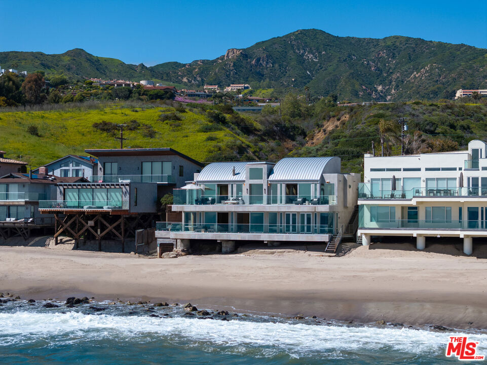 Both a private beachfront retreat and a majestic setting for grand entertaining, this elegant architectural estate is filled with natural light and sensational ocean, island, and coastline views. With approximately 81 feet of beach frontage on prestigious Malibu Road, the residence has been gorgeously updated for comfort, quality, and architectural integrity. Complete with pool, full-width balconies on two levels, walls of glass, and luxurious indoor-outdoor entertaining spaces, this is a year-round getaway convenient to Pepperdine University, beaches, trails, and all the attractions that Malibu has to offer. Gated and private, the home is built around a tranquil ocean-view courtyard, which has an expansive deck, a firepit lounge area, an outdoor kitchen and barbecue, and a waterfall swimming pool and spa. A door from the courtyard provides stairway access to the pristine beach below. The home's bold lines are emphasized by a long, ocean-view hallway with skylights overhead and up-lit glass insets in the floor. Along this hallway is a modernist wet bar with views of both the pool and the ocean, plus bar seating, a stone trough sink, and cabinets and counter space for spirits and storage. The end of the hall opens into two dramatic, oceanfront spaces with soaring barrel-vault ceilings, clerestory windows, and huge glass doors that open to the upper-level deck. On one side, the handsomely furnished living room has a linear fireplace, a built-in display bench, and glass doors opening to both the pool and the deck. On the other side, also opening to the deck, is the impressive kitchen/dining area. The kitchen is spacious both for food preparation and for entertaining. A large island with bar seating has a gorgeous live-edge countertop. Top-quality Miele appliances, generous storage and counter space, including lighted glass-front cabinets, and an over-sink picture window with views of Point Dume will please the most demanding chef or host. Adjoining the kitchen, with another barrel-vaulted ceiling, is the dining area with ample space for large gatherings that can extend easily to the deck just outside. Completing the home's main level is an ocean-view bedroom suite, a laundry room, and a powder room. On the lower level are three bedrooms, all opening to another beachfront deck. The primary suite is a private haven, with beautiful wood finishes, a platform bed, a sitting area separated by a double-sided fireplace, a wet bar, a walk-in closet with a magnificent burl bench, and a romantic spa-style bath with a sauna. In addition to the main deck, the primary suite opens to a pergola-shaded deck at the side of the house, where a hot tub, hammock, and lounge chairs offer a private, ocean-view oasis. A two-car garage, smart-home systems, and generous storage complete this sublime beachfront estate.