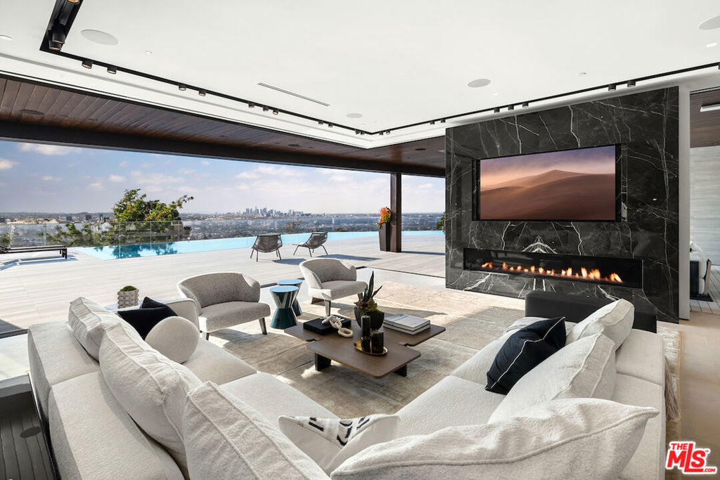 Situated in a prime lower hillside location above the renowned Chateau Marmont, this newly constructed residence offers the epitome of luxury Los Angeles living. Past a double gated motor court, the home's modern steel, stone, and glass facade opens to a truly luxurious interior created by award-winning Nathalie M Designs that is filled with disappearing walls of glass, architectural clean lines, European Oak Wire Brushed floors, 6,399 square feet of deck space, and thoughtful high-end finishes. A functional open layout includes the main living area, grounded by an impressive stone fireplace, and grand dining area, both intended to take in the uninterrupted skyline views. A contemporary dual island kitchen with a secondary dining area sits open to both the main living and dining areas, outfitted with custom two-tone cabinetry, beautiful shadowy stone waterfall surfaces, and seamless top-grade appliances. A designer butler's pantry, guest powder room, secondary ensuite bedroom, and car gallery round out the main level. Sliding glass walls pocket seamlessly from the main living area for uninterrupted access to the outdoor deck and picturesque zero-edge pool with spa, designed with intricate tiles and a relaxing shallow sundeck. Glass railings allow for every opportunity to take in the views, while a covered lounge area and bar-edged outdoor kitchen provide ample entertainment areas. The upper level of the home includes the dazzling primary suite, crafted with a stunning stone-facade fireplace, purposeful wet bar, substantial custom closet, a stone-wrapped spa grade bathroom, and of course, glass pocket walls to access the private patio and take in the incredible views. A plush lounge area, outdoor patio, and three secondary bedrooms all featuring ensuite designer bathrooms and some including private outdoor spaces, complete the top floor of the residence. Accessed via floating staircase from the second level outdoor patio, or interior elevator which is accessible to all floors and rooftop, the pinnacle of the property features an expansive rooftop deck, outfitted with a full outdoor kitchen, gas firepit, and glass railings for an epic vantage point. At the lower level of the home, a secondary living area contrasts sleek white walls with a magnificent book matched stone fireplace and entertainment area, ultimately leading to two additional secondary bedrooms with designer ensuite bathrooms, a plush galaxy-ceiling theater, designated laundry room, and resort-level spa, complete with oversized soaking tub, glass enclosed shower, and stone steam room. Additional aspects of the home include a second garage, intricate lighting to accent key architectural features, a state-of-the-art surround sound system, top of the line smart home technology, and an impressive fully equipped home security system.