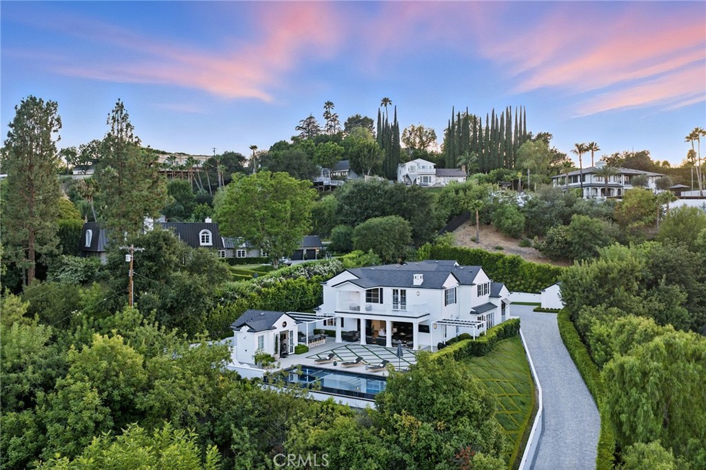 Tucked behind private gates at the end of a long, secluded driveway and surrounded by an enchanting English garden, this modern Cape Cod estate offers a rare blend of privacy, luxury, and timeless style. Perfectly hidden from view and free from outside intrusion, the property is a true retreat from the city. A grand and inviting entry welcomes you into an impressive open-concept layout, where soaring ceilings and walls of glass flood the interiors with natural light. Gleaming wood floors guide you through the home, leading to a light-filled dining room framed by oversized French doors and overlooking a picturesque interior courtyard—an ideal space for intimate gatherings or quiet moments.The gourmet kitchen is a culinary masterpiece, outfitted with top-of-the-line appliances, a double island with seating for informal dining, and a spacious butler’s pantry. Just beyond, a sleek and expansive bar transitions effortlessly into the family room, creating a warm, social hub for entertaining. Oversized pocket doors disappear into the walls, opening the home to a seamless indoor/outdoor flow. Step outside to a private backyard sanctuary boasting unobstructed views, a striking infinity-edge pool and spa, a stylish pool house, and a spacious patio. Enjoy evenings by the fire pit, play on the lush grassy lawn, or host al fresco meals in the covered dining area complete with a built-in BBQ. The luxurious master suite is a true retreat, offering panoramic vistas, a serene sitting area, and a private deck perfect for morning coffee or sunset views. Dual walk-in closets provide abundant storage, while the spa-inspired en-suite bath completes the experience. Ideally located with convenient access to the 101 and 405 freeways, plus discreet back-route access to the Westside, the property balances accessibility with complete seclusion. Designed for both grand-scale entertaining and everyday comfort, this estate is more than a home—it’s a lifestyle