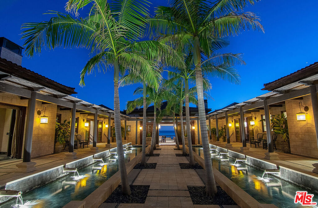 With breathtaking views, Bali-inspired design, and every contemporary comfort, this magnificent estate is a serene sanctuary for exquisite living and impressive entertaining. The private, gated property of over 1.2 acres extends from PCH to Malibu Road, perfectly landscaped and commanding expansive ocean views. A long palm-lined drive leads past a two-story, two-bedroom guesthouse, a lighted north-south tennis court and pavilion, and a fountain, waterfall, and gardens before arriving at the enormous motor court and four-car garage. A dramatic gateway and long, serene palm-lined reflecting pool lead between the wings of the U-shaped house to the gorgeous double-door entry. With interiors designed by Martyn Lawrence Bullard and stunning sight-lines to the ocean, the house is detailed with ornate wood carving, very tall beamed ceilings, wood and stone floors, and full-width exterior doors that vanish to offer seamless flow between indoors and out. The entry steps down to the formal living room, which includes a generous dining area, a pair of facing fireplaces, and a wall of glass that opens onto a roomy patio with a fireplace, stunning ocean view, and a nearby fountain and barbecue island. Adjoining the living room is a spectacular ocean-view great room that includes a family room with fireplace, dining area, and a gourmet kitchen with a huge stone-topped island with seating, deeply recessed tray ceilings, beautiful cabinetry, and a pantry. One wing of the house has two bedrooms and a laundry room. The other wing features a majestic ocean-view owner's retreat with high wood beamed ceilings, beautiful wall coverings, wood floors, and a wide glass doorway opening to a tranquil covered patio. A hallway leads to a luxurious bath, two very large closets/dressing areas, and, at the end of the hall, a resplendent Bali-inspired library/study with high coved ceilings, a wall of built-in book shelves and cabinets, a fireplace, and doors to the patio and to a private sitting area. The home's lower level features a party-perfect bar room highlighted with ornate metalwork that encloses a large wine storage area. The bar extends into a spacious tiered home projection theater. A long hallway leads to a second powder room, a changing room, and another bar, which opens to the lower patio, spa, sitting areas, and lawns, as well as the infinity ozone pool, patio, cabana, and private steps to Malibu Road and access to the beach/ocean. With Crestron electronics and full-house water filtration, this remarkable estate is a livable legacy.