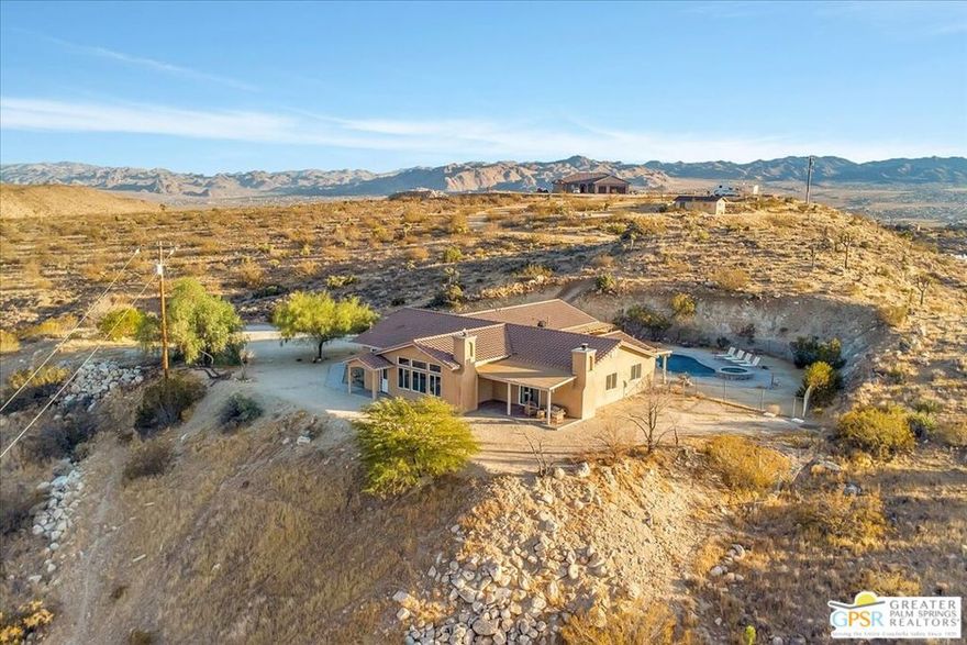 HILLTOP RETREAT on more than 5 acres in Yucca Valley. A private drive leads to this striking architectural property with panoramic views over The Mesa and Flamingo Heights. The private pool and spa with waterfall sparkle by day and come alive at night under the desert stars. Inside, beamed and vaulted ceilings crown the main living areas, where living, dining, and the gourmet kitchen flow seamlessly together. Polished concrete floors add style, while walls of glass showcase endless views. The kitchen is designed for entertaining with two side by side islands, counter seating, and a breakfast area. The primary ensuite features dual walk in closets, a fireplace, dual vanities, separate tub and shower, and direct access to a private patio. Two guest bedrooms each enjoy their own full bath, and a utility room sits along the central hall. Additional highlights include garage parking for three cars, abundant guest parking, and RV parking. The home has also enjoyed a successful vacation rental history, appealing to both investors and part time residents, and is just minutes from Pioneertown, Joshua Tree National Park, and the best of Yucca Valley, combining privacy, views, and desert lifestyle in one remarkable package.