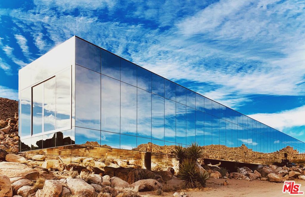 There are only a few spaces in the world that capture architectural brilliance, interior design magnificence, and environmental harmony all at once. Now is the chance to own something truly remarkable, seemingly unattainable. Welcome to the Invisible House, 67.5 acres of pure tranquility in Joshua Tree, California. Designed by film producer, Chris Hanley, and Frank Gehry collaborator, Tomas Osinski, the Invisible House is built to directly connect inhabitants with the environment through a mirrored glass exterior that seamlessly bridges the gap between indoor and outdoor space, masking itself as an indistinguishable component of the dramatic, rocky, desert landscape Joshua Tree is renowned for. But it's not just about how the home functions within its environment, it's also about what it provides for the residental holistic human experience that evokes a feeling of natural peace and wonderment through a truly unique inTruterior design concept that combines the beauty of the desert with luxurious modern function. The conversation piece of the interior 100-foot, HTP solar heated swimming pool running along the west wall, offers a natural indoor element built for pure relaxation, and is equipped with radiant ambient lighting that both illuminates the landscape and communicates with the constellations above. A world-class chef's kitchen features dual ovens, separate refrigerator and freezer, storage, gleaming white marble surfaces, Boffi fixtures, and Miele, Sub Zero, and Wolf appliances all anchored upon sleek and smooth concrete flooring and accented by minimalist lines. Each spacious living area featuring modern Flos lighting is connected to an entire wall of sliding glass doors all opening to the breathtaking landscape, offering a one-of-a-kind living experience that blurs the line between home and nature. Awaken every morning in each of the large three bedrooms to sweeping views of the landscape all equipped with sliding doors, as well as en-suite Boffi bathrooms with a custom all-glass exposed shower in bath #3. A large primary suite offers a private sanctuary and includes a 5-star-resort-inspired bath with soaking tub, floating vanities, and rain shower. For the true visual artist, a massive 224 square foot white wall at the end of the pool is designed for screening films or projecting photographs. The 5,490 square foot living space offers plenty of room for private enjoyment or entertaining, measuring 225 long, by 24'11 wide, with a facade height of 21, and a soaring interior interior height of 12. The remarkable, energy efficient space also features 10,500 of Vitro spectrally selective Solarcool blue glazing with a nominal 12mm airspace, all Title 24 approved. Other modern amenities include 800 amps of electric service; a Vision Coolroof with a 38R value, and closed cell foam insulation; 92 Sunpower 360w, USA-made PV solar panels with Enphase micro inverters all on an IH smart system; 3 Tesla 13kw Powerwall Batteries, and online smart system with solar panels; HTP solar thermal heat panels featuring 20 4 x 10 panels providing 40,000 BTU's each of solar hot water for pool and interior heating; a Halo whole house water filtration system; Starlink high broadband internet with two additional broadband systems including Mojave Wi-Fi and Flashbyte; Orsini HEPA filters for dust, bacteria, and virus eradication, Raypak 27kW pool heater; and two Oxygen ozone pool filters and UV pool filter. Set adjacent to rocks for passive solar heating and cooling and bordered by Section 5 Federal National Park land and a Section 6 BLM, gated nature/tortoise reserve, this once-in-a-lifetime property delivers an idyllic balance of serenity and wonder. Realize the pinnacle of architectural and visual mastery and become one with the environment that surrounds you. The Invisible House offers an unparalleled human experience in one of California's most iconic locales surrounded by a thriving and vibrant community of artists, naturalists, and dreamers