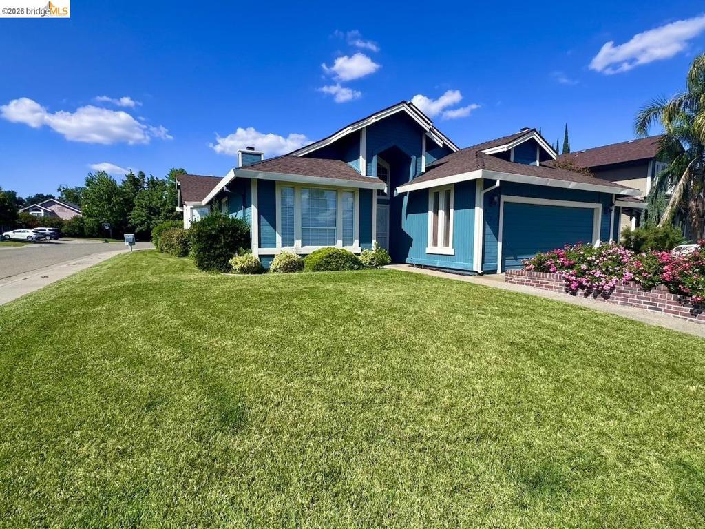 Exterior featuring a vibrant blue siding, white trim accents, a two-car garage, established shrubbery, and a brick-lined flower bed with blooming plants