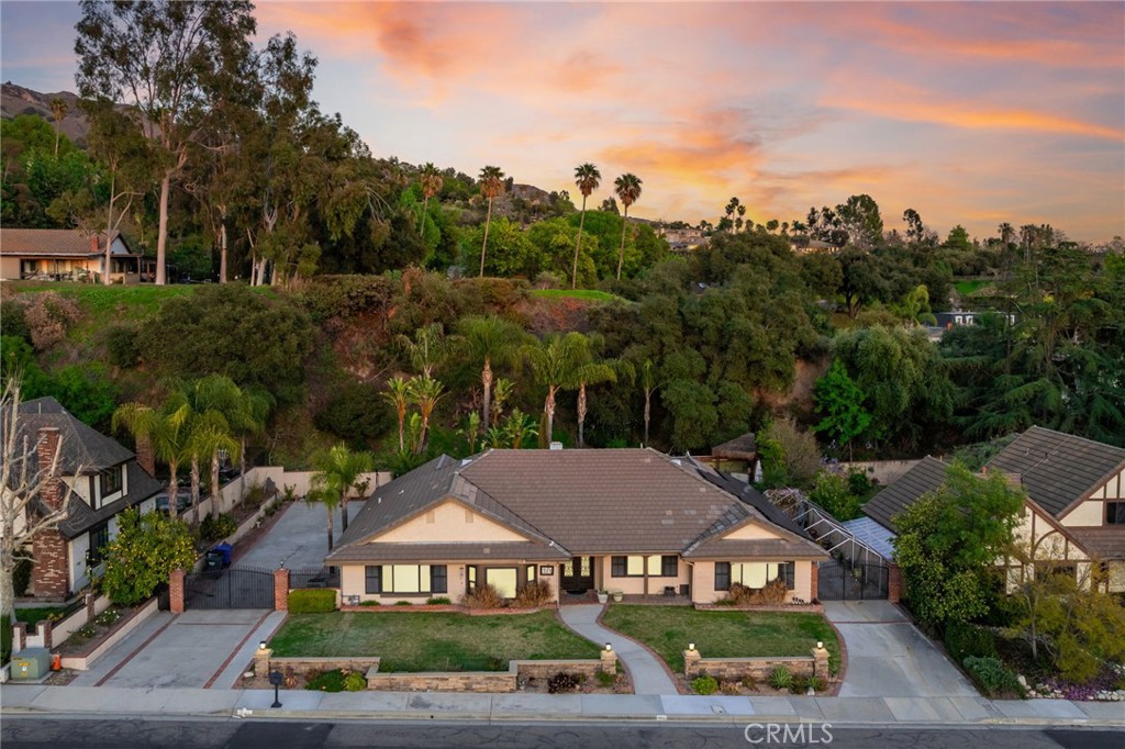 North Glendora Single-Story Retreat Framed by a Hillside Backdrop. The meticulously landscaped front yard features lush lawn, vibrant planters, elegant hardscaping, and a charming brick-lined front porch that creates an inviting first impression. Enter through double iron-and-glass doors into a warm, light-filled entryway where herringbone-patterned wood tile flooring meets a dramatic exposed beam ceiling, blending rustic charm with modern sophistication. At the front of the home, the formal living room offers a serene bay window overlooking the pristine front yard, enhanced by timeless plantation shutters. Just beyond, the formal dining room showcases a wood-detailed tray ceiling and French doors that open to a peaceful side yard. The updated kitchen features granite countertops, stainless steel appliances including a six-burner gas cooktop with a sleek hood, recessed lighting, and a large center island with bar seating. A breakfast nook connects the kitchen to the expansive family room, where vaulted ceilings with exposed beams rise above a floor-to-ceiling brick fireplace. French doors lead out to the covered patio, creating effortless indoor-outdoor flow. The primary suite is a private retreat, complete with a sliding glass door to the backyard, a generous walk-in closet, and a luxurious en-suite bathroom featuring dual sinks, a vanity counter, and a tiled walk-in shower with frameless glass enclosure. Three additional bedrooms offer flexibility for family, guests, or a home office—two with coordinating tile floors, and one highlighted by rich bamboo flooring. The hall bathroom echoes the primary with dual sinks, a vanity counter, and a shower/tub combo. Step into your own resort-style backyard with tropical flair—complete with a sparkling pool and spa with stone coping, a covered palapa, a cozy fire pit, artificial turf lawn, and a built-in barbecue for entertaining. Mature trees and a dramatic hillside backdrop create a peaceful and private setting. Additional features include an automatic double gate to a side-entry two-car garage with ample parking space, plus a second gated driveway with RV parking. A fully owned solar panel system provides energy efficiency and long-term savings.
