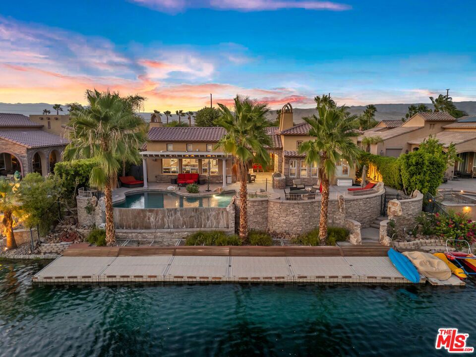 RARE OPPORTUNITY TO OWN ONE OF THE MOST SPECTACULAR AND LARGEST HOMES IN SANTO TOMAS!!! LUXURY LAKE FRONT DESERT LIVING AT ITS FINEST!!! Perfectly situated on a premium SOUTH-FACING lot in the highly desirable gated lake community of Santo Tomas, 64 Via Santo Tomas offers a unique south-facing backyard with plenty of luxurious amenities - infinity pool with a tanning ledge, hot tub, built-in BBQ station, fire pit, fireplace, and steps leading to the massive private dock. Located at the center of the community with unobstructed open water views of the nine acre lake, this home (one of only three) is perfectly situated directly across from the main entrance gate. The 3,350 sqft house boasts four bedrooms and four-and-a-half bathrooms (four are en-suite) with upgraded Kentucky Oak floors, security system, two car garage with plenty of storage space, two indoor fireplaces (one gas and one wood-burning), a bar area, and separation of bedrooms for added privacy. Granite countertops, stainless steel appliances, plenty of cabinets, and a walk-in pantry in the massive kitchen allow for easy entertaining. Two of the bedrooms can be used as an attached casita with a separate entrance door. Home comes fully furnished and has many upgrades - roof (2023), HVAC unit (2024), air conditioned garage, misters in the covered patio, and so much more. Low HOA dues includes a social and sports membership to the Mission Hills Country Club (pool, tennis, fitness, croquet, pickleball, restaurant). Enjoy desert living at its finest!