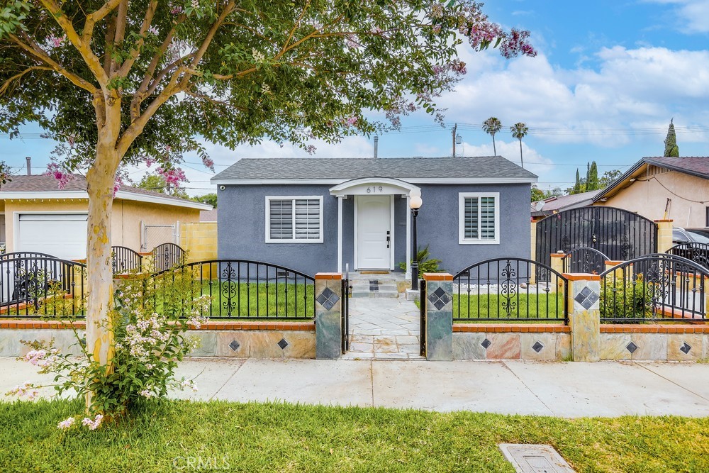 Welcome to 619 S Olive Street, a bright and inviting single-level home in Anaheim offering a spacious lot, private bonus space, and thoughtful indoor-outdoor flow. A stone walkway leads through a quaint, low gate and onto a small front patio framed by a symmetrical exterior—centered front door, windows on each side, and a soft arched overhang that gives the home a classic, almost storybook look. To the right, the path continues alongside the house where fencing opens to a gated driveway leading to the backyard and secondary access to the home. Inside, natural light pours in through windows. Light wood flooring and white interiors add to the bright, airy feel. With the kitchen to the right and a cozy living area to the left, the layout blends comfort and functionality. Off the living room is a bedroom with a walk-in closet and en-suite bathroom featuring a dark wood vanity, crisp white finishes, and a unique tub/shower alcove. The bathroom also connects to a hallway that ties through the kitchen and leads to an additional bedroom with its own closet and convenient access to the rest of the home. Out back, the yard features circular garden beds with established trees that break up the paved space and add greenery to the setting. Multiple gated access points offer flexibility and privacy—ideal for multi-use living or entertaining. Toward the back of the property is a converted garage, now a detached bonus space with its own entrance, main room, full bathroom, and a separate niche for storage or wardrobe use. The unit has its own A/C, making it perfect for a guest suite, office, or studio. Located in a desirable Anaheim neighborhood, 619 S Olive Street offers abundant outdoor space, smart design, and adaptable living in a uniquely flexible and well-connected setting.