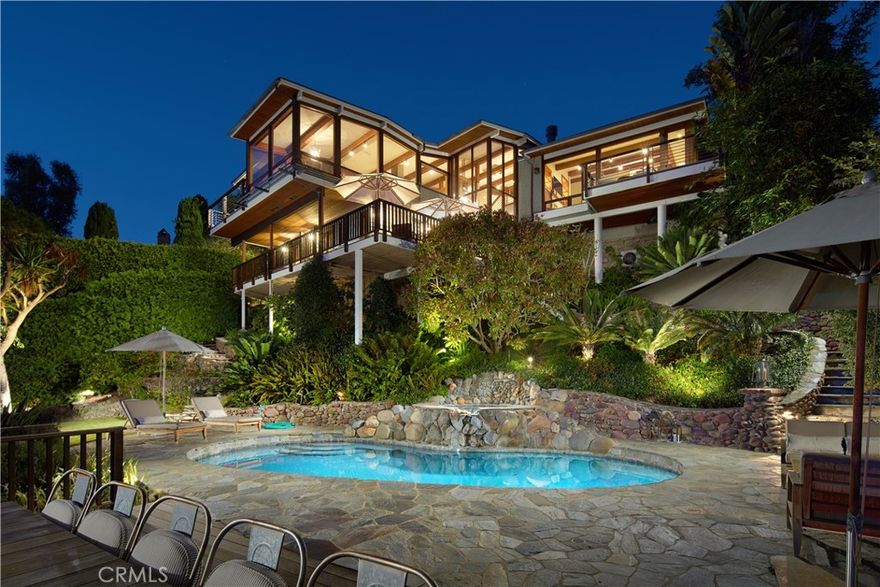 Designed by renowned architect Horst Noppenberger, this breathtaking approx. 3,665-square-foot coastal residence blends warm wood and glass elements with inspired modern design to capture panoramic ocean views and effortless indoor/outdoor living. Spanning three levels, the home welcomes you from the street-level entry with a light-filled foyer featuring clerestory windows, skylights, and custom woodwork, along with interior access to a two-car garage and four additional uncovered parking spaces.

Descending to the main level, expansive living and entertaining spaces unfold—a great room anchored by a stone fireplace and rich custom cabinetry opens completely to covered terraces, creating a seamless connection between the interiors and the outdoors. This level features an ocean-view dining nook wrapped in glass, a well-appointed kitchen with polished Afyon white marble countertops, a media room, a half bath, and the ocean-view primary suite—a serene sanctuary with its own fireplace, sitting area, private balcony, and a luxurious bath with dual sinks, soaking tub, separate shower, and walk-in closet with built-ins.

On the lower level, two oversized ensuite bedrooms and a laundry area provide comfort and flexibility, while stairs lead further down to the lush, terraced backyard centered around a sparkling pool and spa with waterfall feature, multiple seating areas, and a fully equipped outdoor kitchen with bar, grill, and fireplace—perfect for entertaining or sunset gatherings. In total, the home offers approx. 877 square feet of ocean-view decks, enhancing the seamless indoor/outdoor lifestyle.

Thoughtfully appointed with premium finishes and technology, the residence features a Vantage lighting system for sophisticated, customizable illumination throughout.

Tucked into the hills above Woods Cove, this architectural gem offers the best of Laguna Beach living—sweeping ocean and coastline views, proximity to pristine beaches, the Village, and the town’s celebrated art, dining, and coastal culture—all from a quiet, private setting that feels worlds away.