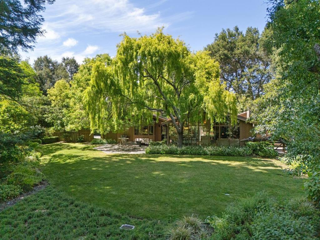 Mid-Century Modern Style on 1.3 Acres Blocks from Town Center. Classic mid-century modern architecture is showcased on beautiful grounds of approximately 1.3 level acres just blocks from Town Center. Complete privacy is offered behind gates, where a long driveway leads past a fenced tennis court to the home and detached garage. Inside, the ambiance is airy and light-filled with soaring wood-paneled ceilings, skylights, and an entire wall of glass that frames the natural surroundings. Beautifully appointed throughout, the home has hardwood floors, dazzling modern lighting, and a sleek open layout with 3 bedrooms, an office, and 2.5 baths. The formal living & dining rooms as well as the kitchen share a dramatic wood-paneled cathedral ceiling and the wall of glass. A fireplace plus floor-to-ceiling library shelves with moveable ladder access are highlights. A separate family room anchors one wing with two bedrooms and a bath, while the primary suite and office are privately positioned in a separate wing, which has access to a patio with large spa. The grounds are a true retreat with redwood groves, creekside sitting areas, raised vegetable beds, tennis court, plus expansive areas for outdoor entertaining. Access to acclaimed Woodside Elementary School (K8) adds the finishing touch.
