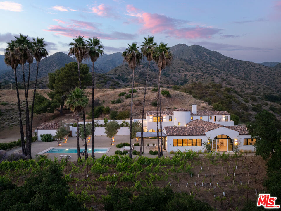 Perched on a private knoll in Ojai's coveted East End, this 2023-built estate blends modern Mediterranean design with refined living. The 5,100 sq ft villa sits on nearly two secluded acres and includes a fully licensed, organically farmed boutique vineyard with over 1,300 mature vines.Thoughtfully crafted with top-tier materials, the home features a dramatic great room with 12-foot ceilings, floor-to-ceiling French doors, groin-vaulted ceilings, and a sculptural circular staircase. White cement floors, hand-plastered walls, and arched passageways add timeless elegance.Designed for both entertaining and everyday luxury, the chef's kitchen boasts Sub-Zero refrigeration, Fisher & Paykel ovens, and Waterworks fixtures, flowing into open dining spaces and a veranda perfect for enjoying Ojai's "Pink Moment" sunsets.Four private bedroom suites, each with spa-style baths and outdoor terraces, provide serene retreats. The grounds offer ancient oaks, an olive allee, drought-tolerant gardens, a heated pool, spa, and multiple outdoor living areas.This estate combines beauty with function. Plans for an ADU, a potential winemaking facility, and safety features like fire-resistant materials, private hydrant, and emergency turnaround ensure long-term peace of mind.A rare blend of design, nature, and opportunitythis East End estate is more than a home; it's a legacy.