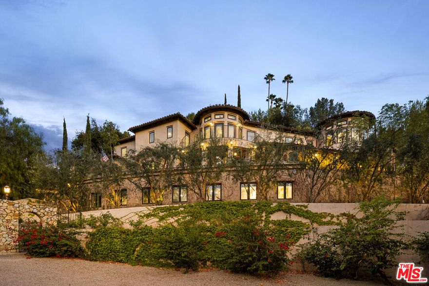 Welcome to this reimagined custom built European Villa with stunning views and magnificent sunsets is set back from the street up an approx 500' drive and set on its own private hilltop knoll. This rural country lane cul-de-sac is the site of many celebrity owned estates and offers a peaceful feeling of living in the countryside yet minutes from legendary Ventura Blvd shopping, restaurants and commuter routes. As you pass through the grand entrance gates, you enter a private world class sanctuary of mature trees set on an incomparable 1.27 acre lot where the possibilities are only limited by your own imagination. This handcrafted original owner's legacy estate welcomes you as you pass the olive grove and arrive to a 25 car motor court that could be the future sight of paddle/tennis courts, sports courts, soccer field and more! An oversized 3-car garage with stone arch accents as well as a multi-car porte-cochere welcomes both the homeowners and their lucky guests. The grand foyer w/ fireplace & soaring ceilings sets the stage for the architectural magnificence that awaits. Hand hewn wide plank solid walnut floors flow through most rooms in the home. A true executive home office will make you never want to leave. The stately art gallery hallway is approx. 120-ft of space that has walls of glass bringing natural light and greenery into view. A banquet-sized formal dining room caters to both large scale events as well as intimate family occasions. The chef's kitchen offers granite counters, stone tile floors and a suite of professional stainless appliances opening to a breakfast room rotunda and the outdoor living dining patio beyond. Additional entertaining spaces include a vaulted ceiling den with fireplace perfect for a library or game room. The adjacent media room is both a family/screening room as well as a bar room area opening to the patio. There are 2 generous sized en suite bedrooms on this level with separate entrances. The penthouse primary suite is truly the crowning glory of the home with an additional art gallery hallway and endless views over the Valley, treetops and mountains beyond. The main bedroom area is flanked by a sitting room/additional office space, dual baths (one w mosaic tile and the other with white Calcutta marble) 2 massive movie star worthy closets and a generous home gym that could be future additional bedroom space. There is an elevator that serves the main and penthouse levels as well as the garage and home office level. The lower level home office space has been the site of a successful business for approx. 17 years and offers approx 1,700 sq ft of limitless options. Future kids fun zone, screening room, additional bedroom space, etc. Currently there is a service kitchen, bathroom and 2 enclosed office spaces with its own entrance. The property even has its own fire hydrant! This one-of-a-kind custom Villa is ready for its next privileged owner to move in and experience what The Good Life truly means. Celebrate your success, good fortune, family & friends at this legendary estate that has been the site of numerous Hollywood parties and adventures and now awaits a new steward to usher in a new era of fame and fortune and tranquility.