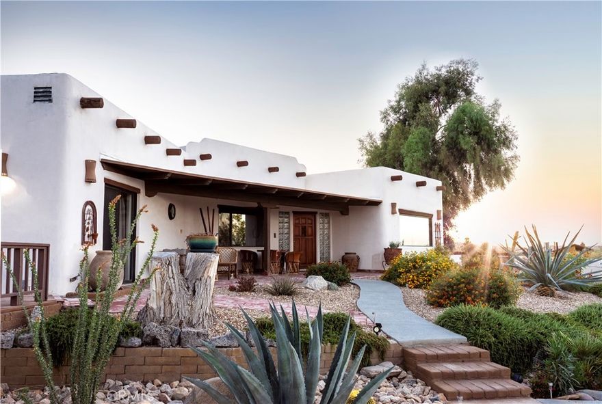 Luxury Spanish Mediterranean Villa With Pool & Fully Furnished! As you travel to this one of a kind property, you’ll be captivated by the stunning backdrop filled with cacti, desert flora, and the majestic Boulder Mountains of Joshua Tree National Park,minutes away. Pulling up to the 2.5 acre estate evokes the feeling of checking into a boutique resort. An architectural gem, this masterfully designed modern Adobe home features boutique aesthetic accents,wood privacy fencing and tastefully executed landscaping that welcomes you in. 
Offering over 2,000 square feet of space, this large 3 bedroom 2 bathroom home, encompasses the definition of a “unique” and open floor plan! The well-appointed foyer flows effortlessly into the rest of the living area, boasting high ceilings, craftsman modern Adobe-style finishes, vaulted open beam ceilings, tasteful built-in shelving, beautiful wood laminate flooring, and a custom-designed fireplace that centers the room. 
The large gourmet kitchen features stainless steel appliances, plenty of kitchen cabinets, fresh design elements, and Santa Fe countertops/backsplash! Just off the kitchen,find the formal dining room, perfect for holiday dinners or any type of gathering! Both guest bedrooms offer ample space, trendy wood laminate flooring, generous closets, and view windows. The full guest bathroom is outfitted with a remodeled walk-in shower, a modern vanity, and mosaic tile flooring! 
Stepping into the primary suite, you discover the perfect retreat at the end of the day, rivaling that of a boutique hotel. This area features more open beam ceilings, a custom light fixture, and a large primary bathroom showcasing a stunning door, dual vanity sinks, custom mirrors, and a sizable walk-in tiled shower.
the backyard, you’ll find your own private luxury oasis filled with cacti, desert flora, and other  plant life, accompanied by walkways and Mediterranean fencing. Tucked away from the world is the large pool and Jacuzzi; as you take in the view, you’ll be tempted to jump right in and take a dip! Enjoy a soft desert breeze on the rustic covered patio, relax on the large wood deck , or take a nap in the stylish cabana—this is outdoor living at its best!
Just when you think it can’t get any better, it does! Stepping into the oversized masonry two-car garage, currently being used as a game room, you’ll find the perfect space for additional storage, a home jam area, an art studio, or, of course, to house your toys and cars!
