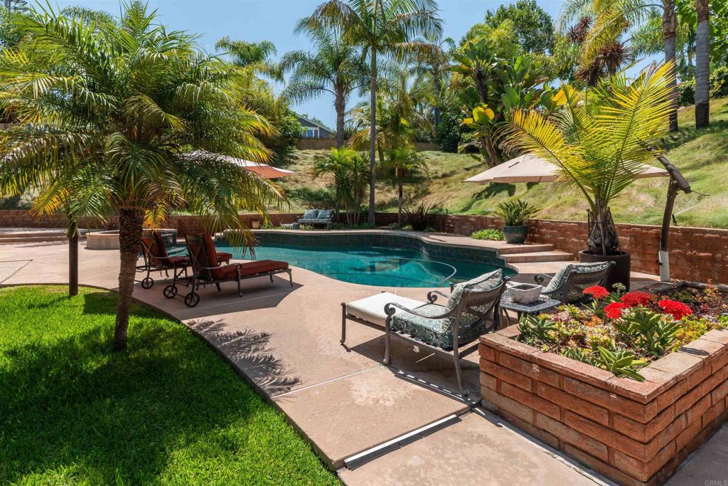 Set on a premier 0.79-acre lot at the end of a quiet La Costa Meadows cul-de-sac, this custom estate blends rare privacy, a main-level primary suite, and sunset views with everyday convenience—minutes from top-rated schools, Alga Norte Park, La Costa Resort & Spa, Aviara Golf Club, shopping, dining, and the coast. Brick entry pillars frame an extra-long paver driveway where layered, drought-tolerant landscaping softens the hillside and opens to a broad motor court. With timeless Mediterranean lines and wrought-iron balcony rails, the home’s curb appeal feels effortless, complemented by a four-car garage that includes an extra-tall bay with lofted storage for gear, hobbies, or a workshop. Terraced steps ascend through the gardens to a glass front door. Inside, the foyer opens with volume and light—vaulted ceilings, a skylit landing, and Brazilian hardwoods that carry through the main level. Just off the entry, the split-level living room gathers around a carved stone fireplace and wide picture windows; three steps up, the formal dining room sits under a statement chandelier, overlooking the living room for easy, connected entertaining. At the heart of the home, the kitchen and family room flow together for everyday ease. Granite counters, culinary-grade built-ins including a six-burner gas range and a central island create a true cook’s space. The adjacent sunken family room adds an inviting second lounge, and oversized sliders open wide to the patio so gatherings flow naturally from the interior outdoors. The main-level vaulted primary suite lives like a quiet hideaway —centered by a corner fireplace and anchored by sliding glass doors that open straight to the pool terrace.The ensuite offers a long double vanity with stone counters, a soaking tub and an oversized glass shower outfitted with multiple sprays and a built-in bench, with a custom walk-in closet behind a solid hardwood door. Also on this level: a chic half bath, a skylit private office, and a tucked-away bonus room ready for a gym, studio, or speakeasy. Upstairs, three comfortable bedrooms and two full baths offer flexibility for family or guests. Two balconies capture long views; one bedroom opens through sliding doors to a view deck. Outside, the yard reads like a resort. Multiple patios wrap the home with a wrought-iron fence framing the upper terrace, and a free-form pool with raised spa and spillway. Brick planters, mature palms, and open sky set an easygoing backdrop for weekend hangs and sunset swims. Thoughtful extras include owned solar, one garage with an extra-tall bay with lofted storage for gear, hobbies, or a workshop, no HOA or Mello-Roos, and a generous, guest-friendly driveway. This is North County living made effortless.