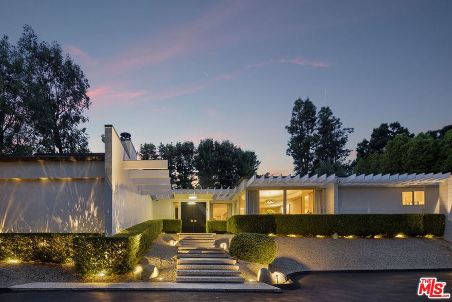 Set behind gates on one of Encino's premier streets, this Mid-Century Modern estate blends iconic architecture with chic, updated interiors. A striking entry opens to a light-filled floor plan accented by expansive glass walls, designer details, and sweeping views of the lush grounds. Elegant living and dining areas showcase custom features, dramatic lighting, and effortless indoor-outdoor flow designed for entertaining. The gourmet kitchen is equipped with top-of-the-line appliances and built-in breakfast seating, while multiple lounge areas provide both style and comfort. Outside, the private backyard retreat boasts a shimmering pool, sun-drenched terraces, cozy fireplace, and mature landscaping that ensures complete privacy. Single-level living at its finest, this residence embodies timeless design and modern sophistication in one of the Valley's most desirable enclaves.