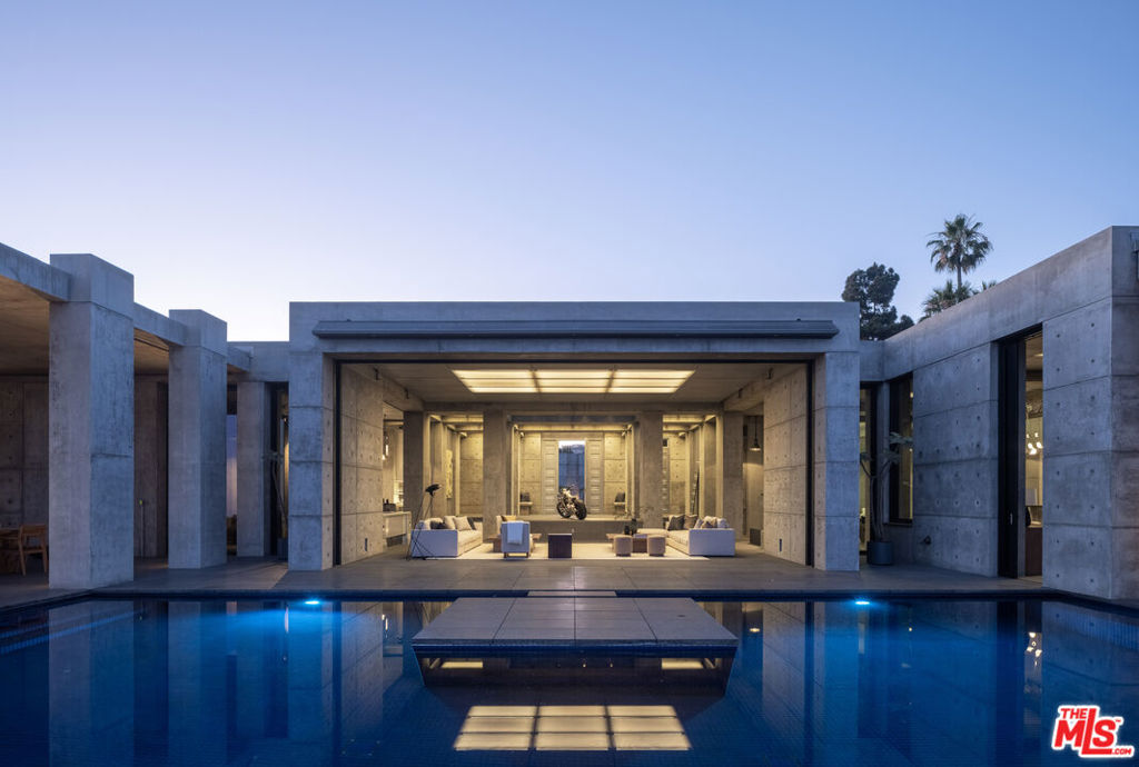 Set at the top of Trousdale, this unparalleled estate is a bold exploration of raw precision, industrial elegance, and sculptural architecture, all while capturing sweeping 180-degree southern views. Designed and built by the acclaimed ID Group, the home centers around a circular motor court enveloped by rhythmic board-formed concrete. Monolithic walls serve as both privacy buffers and dynamic light sculptures, casting dancing shadows from sunrise to dusk. Inside, the architectural intent continues with oversized concrete volumes, perforated metal ceilings, and brushed steel detailing. Framed views of the city and ocean command attention, most dramatically from the main living room, where a retractable glass wall opens to an infinity-edge pool and a horizon that stretches across the Los Angeles skyline. One of the home's most striking features, a fully exposed concept motorcycle, is installed beneath a skylit entry, serving as a dramatic showpiece. Skylights and deep architectural reveals filter daylight through the interiors, creating a constantly shifting interplay of light and shadow. Resort-caliber amenities include a state-of-the-art theater, a spa-inspired primary bath, manicured grounds, and meticulously curated one-of-a-kind fixtures. Every element of the home, from its structural form to its bespoke finishes, has been purposefully designed, making 410 Trousdale a truly singular modernist retreat in the heart of Beverly Hills.