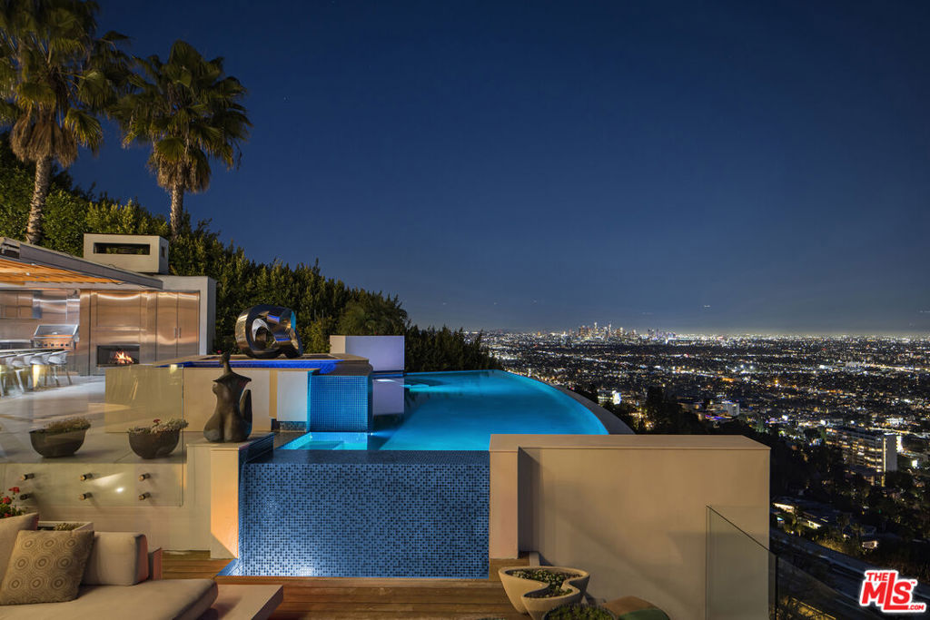 Perched on a rare promontory within a coveted celebrity enclave in The Bird Streets, this extraordinary estate commands a breathtaking panorama, where the city skyline unfolds seamlessly into the Pacific. A cinematic setting once featured in HBO's Entourage, set behind a private gated entry with serene water features, the home is an architectural statement of height and light, seamlessly blending its elevated design with a commanding vantage point over the city.An elegant glass pivot door reveals a soaring double-height foyer, where a glass-bottom floor and floating staircase establish a striking interplay of transparency and light. The formal dining room captivates with floor-to-ceiling awning windows that perfectly frame the skyline, while the expansive living room unfolds onto a sweeping terrace above the clouds, designed for both grand-scale dining and relaxed lounging. A more intimate piano lounge, centered around a fireplace and sleek bar, creates an inviting setting for evening gatherings, while a nearby TV nook offers a cozy retreat, balancing the openness of the main living spaces with a sense of warmth and intimacy. The chef's kitchen, equipped with Miele appliances, large marble island, and espresso bar, captures mesmerizing south-facing views stretching from West Hollywood to Long Beach. A full butler's pantry enhances both function and storage, discreetly serving the culinary space.Ascending to the second floor, the primary suite unfolds as a private sanctuary, complete with an executive office (with full bath and walk-in closet), along with a bedroom fireplace and sliding walls of glass that open onto an extended private terrace with a fitness area. The ensuite is a spa-like retreat, boasting an oversized steam shower with a soaking tub set within, dual vanities, a bidet, and a boutique-style walk-in closet. A secondary primary suite mirrors this level of luxury, featuring a Juliette balcony, dual-sink ensuite with a soaking tub and steam shower, and a generous walk-in closet. The lower level seamlessly integrates entertainment and leisure, with a private guest suite opening onto an outdoor patio with city views. A sleek bar and lounge lead to a sunken theater that transforms into an indoor-outdoor space, perfect for game day gatherings.Outside, the estate's grounds offer a breathtaking extension of the living space. A Viking-equipped outdoor kitchen, complete with a built-in fireplace, sits beneath a covered patio with heating, where an elongated marble dining table accommodates twenty guests. The infinity-edge pool and elevated spa trace the curvature of the property, punctuated by an intricate art installation that plays against the skyline. A mirrored bar reflects the twinkling city lights, complementing an array of luxe amenitiesincluding a craft beer dispenser, sauna, outdoor shower, and full bath. The sunken fire pit lounge, designed to give the sensation of floating above the city, is a showstopping finale to an already incomparable setting.Additional highlights include a three-car garage with private street parking and a unique land extension that reaches Wetherly Drive, offering the potential to establish a more direct access route to Sunset Boulevard. A true rarity in the Bird Streets, this estate is a fully immersive experience in panoramic luxury, where every vantage point is a masterpiece.