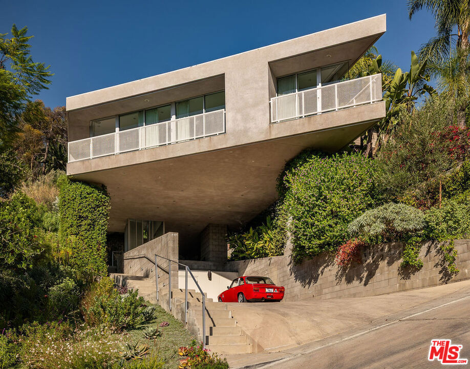 A striking lakeside masterpiece, the House on Six Legs represents a five-year project completed in 2010 by Escher GuneWardena, internationally esteemed architects and preservationists known for their work restoring the famed homes of John Lautner, Paul Williams, Rudolph Schindler, and Charles and Ray Eames. The commanding cantilevered home features 4 bedrooms and 3 bathrooms spanning 2,911 sq. ft. with effortless indoor-outdoor flow to the curated backyard space. Just moments from the Silver Lake Reservoir, this Lautner-inspired residence stands among Silver Lake's celebrated modernist homes. Located at the end of a cul-de-sac, the remarkable home welcomes you with its angular concrete piers and jutting front facade, intentionally built to maximize views and privacy. The cantilevered home extends over the carport, offering a dramatic welcome. Drought-tolerant plantings abut the concrete stairs that ascend to the home's enclosed, front glass atrium. Bordered with floor to ceiling windows and windowed doors, the main living space allows abundant natural light and unobstructed city and hillside views. Dark wood floors contrast with crisp white finishes, creating a bold yet inviting palette. The kitchen serves as both a functional workspace and a gathering hub, with a large-scale island, streamlined cabinetry with hidden storage, and high-end appliances, including Sub-Zero, Wolf, and Bosch. This culinary space seamlessly transitions into dining and lounge areas, allowing a cozy and serene atmosphere. The private quarters continue the theme of light and flexibility. The primary suite showcases a moveable wall that reveals nightly sunsets, a walk-in closet, and a spa-like bath with luxury fixtures by Phillipe Starck, AXOR, and Ronbow. Two secondary bedrooms share a skylit bath designed to maximize brightness, while the fourth room adapts seamlessly as an office or guest retreat, complete with a custom foldaway bed and an integrated workstation. Every room is thoughtfully appointed, with custom window treatments and sleek finishes throughout. The well-kept grounds extend the living experience outdoors, with native plantings, citrus trees, and multiple terraces for entertaining. An elevated deck, built-in barbecue, firepit area, and outdoor dining area create a perfect setting for gatherings large or small, while additional seating areas invite quiet moments under the California sun. Practical features, such as a tankless water heater, automatic irrigation, and a generous four-car parking area with extra storage, add convenience to the commanding architectural form. Located minutes from Silver Lake Reservoir and its premier commercial offerings like LaMill Coffee and Botanica Restaurant, the House on Six Legs offers a unique opportunity for its next owner to be the keeper of this iconic house in the heart of LA's esteemed architectural community.