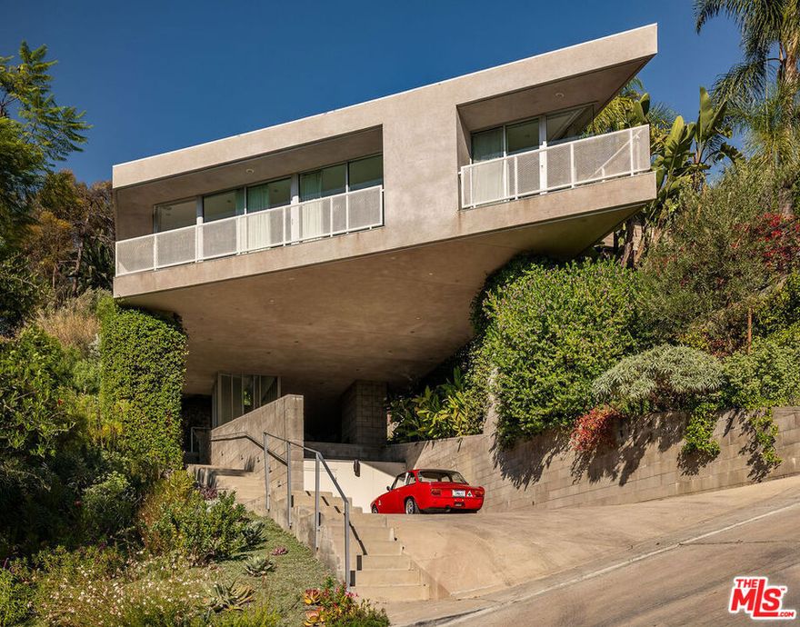 A striking lakeside masterpiece, the House on Six Legs represents a five-year project completed in 2010 by Escher GuneWardena, internationally esteemed architects and preservationists known for their work restoring the famed homes of John Lautner, Paul Williams, Rudolph Schindler, and Charles and Ray Eames. The commanding cantilevered home features 4 bedrooms and 3 bathrooms spanning 2,911 sq. ft. with effortless indoor-outdoor flow to the curated backyard space. Just moments from the Silver Lake Reservoir, this Lautner-inspired residence stands among Silver Lake's celebrated modernist homes. Located at the end of a cul-de-sac, the remarkable home welcomes you with its angular concrete piers and jutting front facade, intentionally built to maximize views and privacy. The cantilevered home extends over the carport, offering a dramatic welcome. Drought-tolerant plantings abut the concrete stairs that ascend to the home's enclosed, front glass atrium. Bordered with floor to ceiling windows and windowed doors, the main living space allows abundant natural light and unobstructed city and hillside views. Dark wood floors contrast with crisp white finishes, creating a bold yet inviting palette. The kitchen serves as both a functional workspace and a gathering hub, with a large-scale island, streamlined cabinetry with hidden storage, and high-end appliances, including Sub-Zero, Wolf, and Bosch. This culinary space seamlessly transitions into dining and lounge areas, allowing a cozy and serene atmosphere. The private quarters continue the theme of light and flexibility. The primary suite showcases a moveable wall that reveals nightly sunsets, a walk-in closet, and a spa-like bath with luxury fixtures by Phillipe Starck, AXOR, and Ronbow. Two secondary bedrooms share a skylit bath designed to maximize brightness, while the fourth room adapts seamlessly as an office or guest retreat, complete with a custom foldaway bed and an integrated workstation. Every room is thoughtfully appointed, with custom window treatments and sleek finishes throughout. The well-kept grounds extend the living experience outdoors, with native plantings, citrus trees, and multiple terraces for entertaining. An elevated deck, built-in barbecue, firepit area, and outdoor dining area create a perfect setting for gatherings large or small, while additional seating areas invite quiet moments under the California sun. Practical features, such as a tankless water heater, automatic irrigation, and a generous four-car parking area with extra storage, add convenience to the commanding architectural form. Located minutes from Silver Lake Reservoir and its premier commercial offerings like LaMill Coffee and Botanica Restaurant, the House on Six Legs offers a unique opportunity for its next owner to be the keeper of this iconic house in the heart of LA's esteemed architectural community.