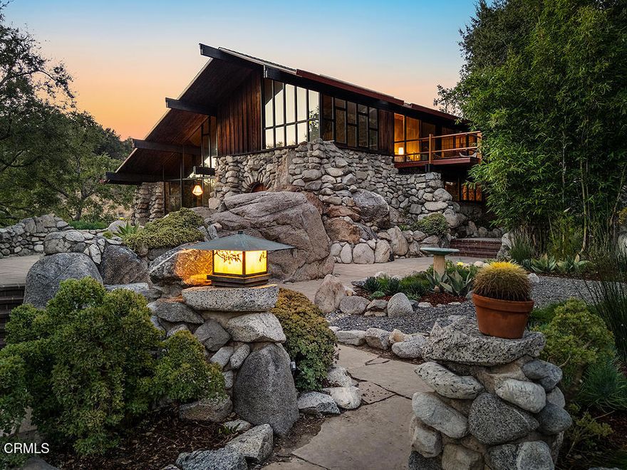 Up a secluded private road in a grove of California live oaks is a creative haven with a unique legacy. 'Boulder Crest' is singular: a historic marvel, an architectural gem, a property rising from the landscape -- an enduring biophilic design. Built of massive granite boulders placed at the beginning of the 20th century, the house and grounds are a feat of vision, engineering and artisan masonry. This home seamlessly melds arts and crafts elements with mid-century modernism - the net effect is timeless - from arroyo stone work and old growth redwood to post & beam construction.  Serpentine stone-lined pathways cross a network of masonry channels and terraces on the approach to the entry. A keystone engraved '1912' heralds over the arched double door, opening to reveal rich jade-hued concrete floors. Redwood paneling, vaulted beamed ceilings and oversize pendant lanterns warm a jaw-dropping step-down living room, anchored by the floor-to-ceiling stone fireplace and wall of windows. The living room connects to both the dining room and galley kitchen with rock walls and clerestory windows, as well as the first bedroom and adjacent den (once a gold vault, ready to be reimagined as a music room, meditation space, or quiet office). Even the 3/4 bath boasts a beautiful river rock wall that imparts a feeling of warmth and security. Upstairs, a spacious art studio enjoys canyon vistas, a private patio and calming sounds of the corten steel water feature. Past shoji screens with encapsulated botanical elements, find the generous primary suite, with its own wall of windows, large cantilevered deck with treetop and pool views, and impeccable full bath. Adjacent, a lofted library space overlooks the living room, separate from the public space, but connected by the beautiful curved stone staircase. The 2.75 acre grounds comprise a large private pool, subterranean workshop, wine cellar, patios, two car garage and over 160 oak trees on two parcels (one measuring 69,006 SF and a second, completely separate lot measuring 50,625 SF). The property sits adjacent to El Prieto Canyon (famed burial place of abolitionist Owen Brown), and at the base of Millard Canyon - in winter, the stream flowing through Angeles National Forest can be heard from the yard. This rare historic home has had just three owners (artists or visionaries, all) -- recognized by Altadena Heritage and published a handful of times, it remains largely under the radar and relaxed.  What a joy it is to behold.