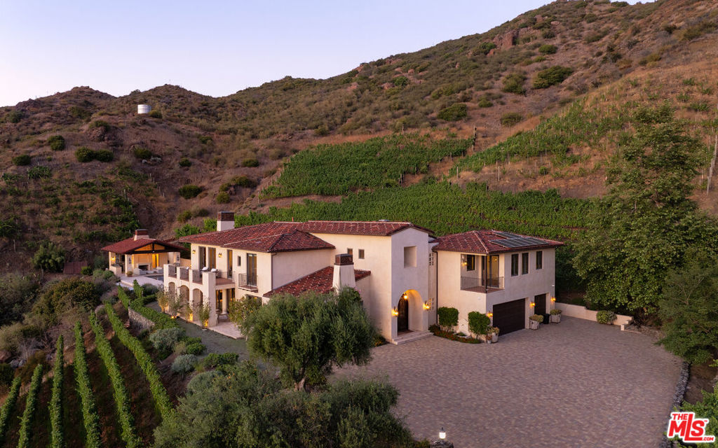 Nestled in the rolling hills of Malibu wine country, 33330 Hassted Drive is a one-of-a-kind vineyard estate where refined living meets natural beauty. Set on a hillside surrounded by mature vines and breathtaking mountain views, this gated retreat offers complete privacy and timeless Mediterranean charm.The main residence is thoughtfully designed with warm stucco exteriors, red-tile roofing, and hand-finished details that echo the rustic elegance of the European countryside. Inside, soaring ceilings, arched hallways, and carved stone fireplaces create a sense of understated luxury. Expansive windows and French doors flood the home with light and frame sweeping vistas of the Santa Monica Mountains from nearly every room.The chef's kitchen is both beautiful and functional, featuring dual center islands, premium appliances, and an open layout ideal for entertaining. Living areas flow effortlessly outdoors to a spectacular pool terrace lined with lounge seating and anchored by a resort-style cabana. Complete with its own fireplace, bar, and shaded dining space, the cabana is designed for unforgettable gatherings from day to night.As the sun sets, the estate transforms into a private cinematic experience with a custom, oversized outdoor movie screen that rises seamlessly from the groundoffering open-air screenings under the stars with the surrounding vineyard as your backdrop. For a more immersive experience, the home also features a dedicated indoor theater room, creating a true dual-screen lifestyle for both indoor comfort and outdoor drama.The primary suite offers a peaceful sanctuary with a private balcony, cozy fireplace, and spa-like bath. A freestanding clawfoot tub is perfectly positioned to take in the surrounding ridgelines, while dual vanities and a large walk-in dressing room with custom cabinetry provide elevated functionality and style.Additional spaces include a handsome wood-paneled office with built-in library shelving and French doors leading to a private patio. A detached guest house offers beautiful accommodations for visitors or staff, while a spacious motor court and three-car garage provide ample parking. Modern comforts are integrated throughout, including a solar panel system and smart home features.From its walkable vineyard rows and inviting interiors to its resort-style amenities and rare cinematic touches, 33330 Hassted Drive is a fully realized Malibu retreat designed for inspired living and unforgettable entertaining.