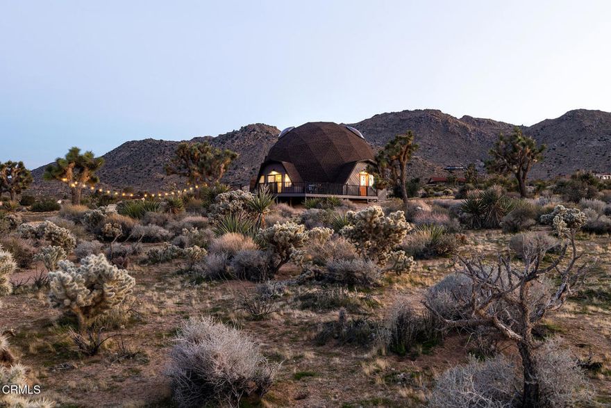 This architectural jewel sits amidst Joshua Trees on over two acres in Upper Friendly Hills. 7677 Wilton Rd is a striking Buckminster Fuller-inspired geodesic dome with soaring ceilings, dramatic skylights, and stunning views of the surrounding mountains and desert. The lower level welcomes you with bamboo flooring and a bright, open layout that flows between the living, dining, and kitchen areas. The kitchen features colorful accents and stainless steel appliances, while the spacious primary suite offers a generous bedroom, large closets, and an en-suite bath. A spiral staircase leads to the lofted second level, where two additional bedrooms with unique triangular windows overlook the grounds. A modern bath showcases designer tile and dual sinks, and a flexible bonus room provides extra living space. Designed to capture natural light and frame the desert sky, the dome's wooden hexagonal details and abundant skylights create a uniquely beautiful atmosphere. Outdoors, the property offers a wraparound deck for al fresco dining, a propane fire pit, and a hot tub perfect for stargazing. With thoughtful upgrades including a newer roof, windows and sliding doors, an updated AC condenser, refreshed baths, and a newer hot water heater, this extraordinary home blends architectural wonder with modern comfort. Whether envisioned as a serene family retreat or a one-of-a-kind vacation escape, this desert dome presents the rare opportunity to live inside a work of art.