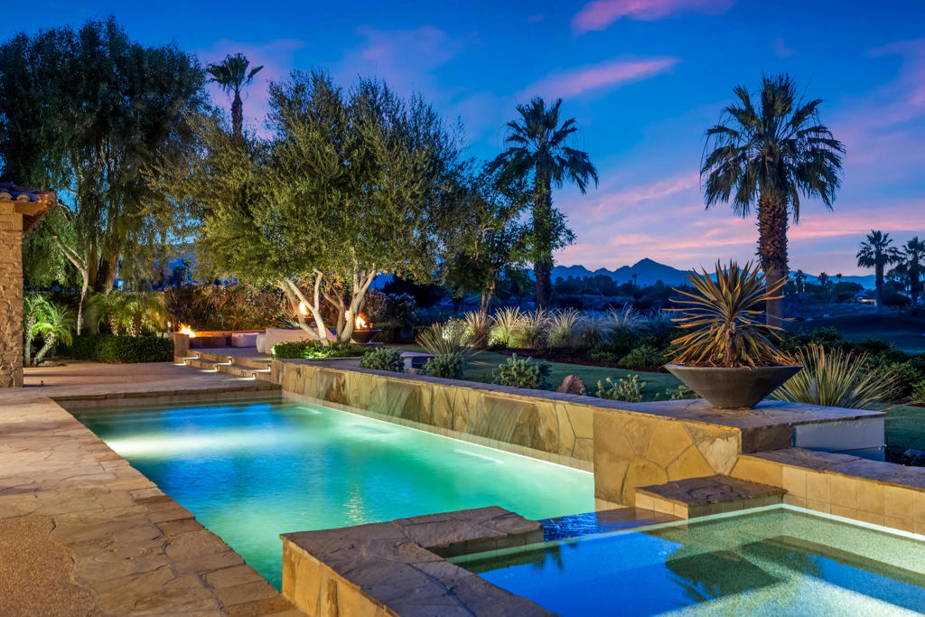 Located behind the prestigious gates of The Hideaway, this custom estate is a breathtaking expression of refined desert living. Blending timeless architecture with contemporary sophistication, it offers a rare opportunity to own a true legacy residence. Set on a premier .37 acre lot with panoramic mountain views and commanding double fairway frontage on the 5th and 8th holes of the Clive Clark course, this 5,266 SF home exemplifies effortless luxury. A gated courtyard opens to a soaring two-story foyer, setting the tone for the elegance that follows. Every element--from scale and proportion to materiality--has been masterfully orchestrated. The great room, anchored by a hand-carved stone fireplace, flows seamlessly into formal and informal living areas designed for entertaining and day-to-day ease. A custom-designed bar with bifold iron doors opens to the covered loggia, creating true indoor-outdoor connectivity. Nearby, the formal dining room and integrated media spaces offer intimate sophistication with a relaxed sensibility. At the heart of the home, the statement kitchen serves as both culinary stage and social hub. A central island, counter bar, custom banquette seating, and a sun-drenched breakfast nook combine form and function. A bespoke iron Dutch door adds charm and practicality, offering easy outdoor access for you and your four-legged companion. Craftsmanship shines through every detail--custom ironwork, artisan stone finishes, tailored lighting, and Waterworks fixtures throughout. The primary suite is a private retreat, opening to two serene outdoor living areas. A custom stone marble fireplace brings warmth and understated elegance, while the spa-inspired bath features a freestanding Waterworks soaking tub, an expansive glass shower with exposed system, and exquisite stonework. Outdoor living unfolds across four distinct spaces: A grand loggia with fireplace and integrated media center A second alfresco dining loggia with fireplace and full outdoor kitchen A main covered loggia as well as a secluded garden retreat exclusive to the primary suite.Additional features include a separate-entry casita, spacious guest suites with private baths and a wet bar, and a resort-style pool with cascading spa, sculptural fire feature, and dramatic fire bowls--set within mature landscaping and rich wood and stone finishes that blur the line between indoors and out. Approx. 5,266 under A/C & Approx. 5,989 SF under roof. Offered professionally furn.