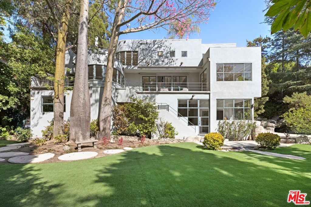 This 1937 Streamline Moderne estate in Los Feliz Oaks offers a rare blend of privacy, scale, and design, set on a 20,415-square-foot lot wrapped in mature trees. Curved walls recall the deck of a ship, lifting the architecture into motion, while inside, double-height ceilings flood the living room with light and a soaring 20-foot fireplace creates a natural gathering point for evenings that linger. Skylights soften the sun, and the kitchen is equipped with a SubZero refrigerator, Viking range, and a breakfast nook overlooking the pond. It shifts easily from quiet morning rituals to holiday dinners filled with friends and conversation. Upstairs, the primary suite opens to a balcony suspended above the city. Downstairs, a screening room, wet bar, and two bedrooms extend seamlessly to the pool, a private retreat with waterfalls, grotto spa, and a wide lawn that welcomes gatherings of every kind. Designed for entertaining yet grounded in quiet retreat, the home balances connection and escape. Close to Griffith Park trails and Los Feliz cafes, it feels both tucked away and within reach. Interior square footage was taped at 4,804 while public record reflects 2,888, buyer to verify.