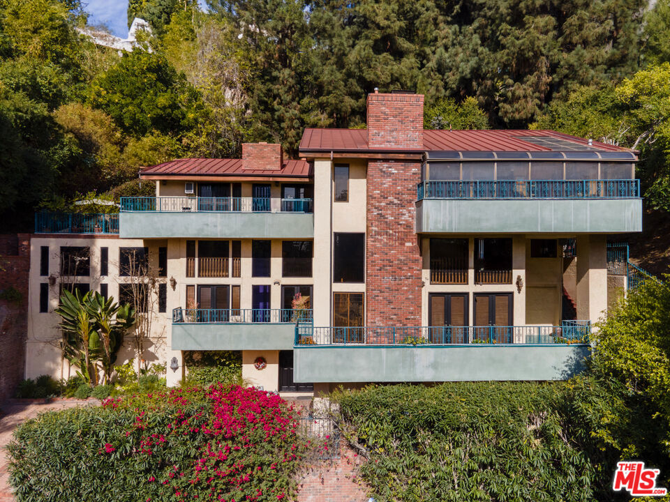 Offered for the first time in over three decades, this grand four-story Traditional estate sits atop a gated 13,611 sq ft lot in one of the most coveted hillside neighborhoods of Beverly Hills/Los Angeles. Encompassing approximately 5,300 sq ft, this 5-bedroom, 5-bath residence seamlessly blends timeless design with sweeping canyon and city views from nearly every room. Thoughtfully positioned on the hillside, the home is bathed in natural light through expansive windows and skylights, including one above the kitchen. Original oak floors, leaded and stained glass, marble fireplaces, and detailed craftsmanship add to the home's refined elegance. The formal living room features a marble fireplace and a wet bar, opening into a solarium with panoramic canyon and city views. Additional highlights include an oak-paneled library with fireplace, a formal dining room with beveled glass, and a gourmet kitchen equipped with granite countertops, a Sub-Zero refrigerator, Wolf double ovens, Bosch dishwasher, center island, and a skylit breakfast nook adorned with stained glass accents. The two-story primary suite is a true sanctuary, offering a private library/office, walk-in closets, a spa-style bath, and abundant natural light. A private, winding staircase leads to the upper level, creating a serene and versatile space perfect for an additional office, reading room, or creative retreat. Additional features include a paneled office or bedroom with its own private entrance and exit, a waiting area, and a full bath. This flexible space can be converted back into a guest suite with a small kitchen area if desired. Ideal for visitors, staff, or extended stays. A game room-style den, extra office or bedroom space, multiple full and half baths, a formal library room, wine cellar, laundry room, elevator, intercom system, and central vacuum. The home features a gated two-car garage plus a carport with space for at least three additional vehicles. Outdoor areas boast a spacious terrace with a dining area, surrounded by mature landscaping and enclosed by a 1030 ft brick privacy wall.