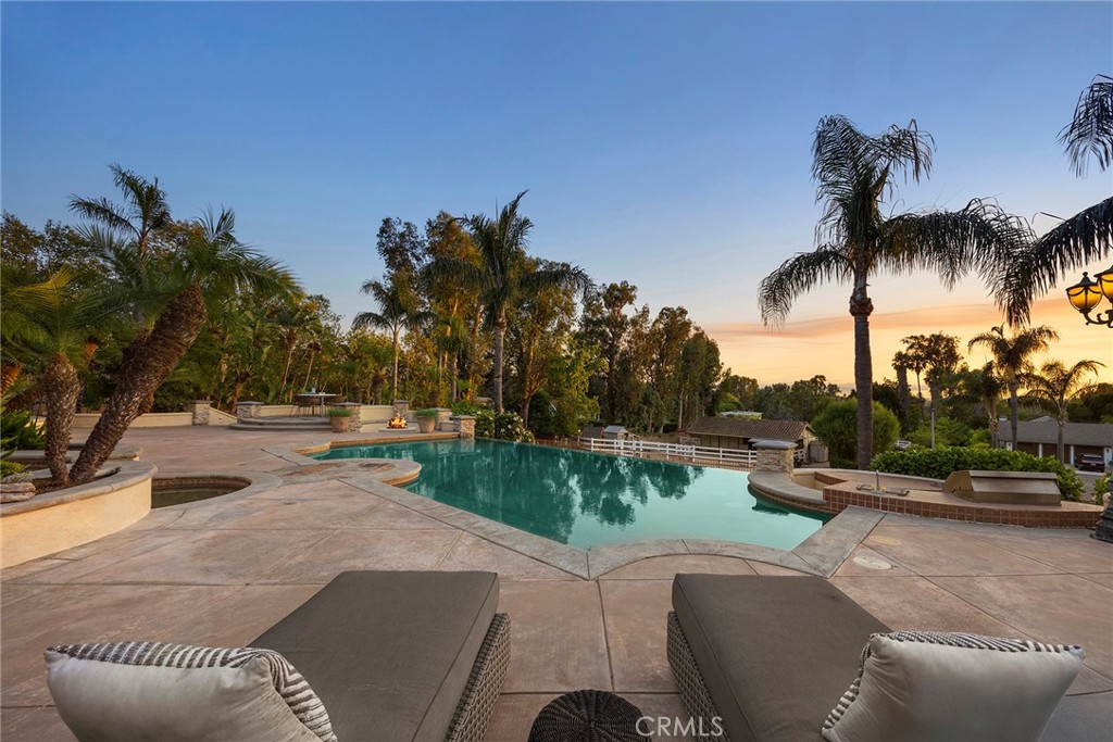 It’s often said that no home is perfect—but this extraordinary estate proves otherwise. Nestled at the end of a serene cul-de-sac behind private gates, this custom single-story residence redefines luxury living with an effortless blend of sophistication, comfort, and unparalleled design. Every detail has been masterfully curated to offer an elevated lifestyle beyond compare. From the moment you step through the grand foyer, you are greeted by soaring ceilings, expansive walls of glass, and a flood of natural light. With sweeping sunset vistas and nearly a full acre of impeccably landscaped, fully usable grounds, this home offers a rare sense of privacy, tranquility & space. The open-concept layout flows gracefully from the formal living and dining areas into a gourmet kitchen and an expansive family room, designed for both grand entertaining and intimate gatherings. Two elegant fireplaces enhance the ambiance, while 10-foot sliding glass doors open wide to create a seamless indoor/outdoor living experience that is simply breathtaking. Designed with thoughtful separation and privacy in mind, the home features three distinct bedroom wings and a private office. The primary suite is a sanctuary overlooking the estate’s most spectacular views. Complete with a fireplace and lavish spa-inspired bath boasting a walk-in open shower, dual vanities, and two custom-designed closets—what an indulgent retreat! Upstairs, a hidden gem awaits—a versatile game room with its own wet bar, built-in entertainment center, and full bath, perfect for relaxing or entertaining in style. Outside, the resort-caliber amenities are endless. A stunning infinity-edge pool with a swim-up bar anchors the outdoor space, complemented by a built-in BBQ island, a bubbling spa, and expansive sun-drenched patios. As evening falls, gather around the fire pit under a starlit sky and revel in the atmosphere of your own private five-star resort. Rolling green lawns, vibrant landscaping, and thoughtfully placed planters lead you to a secluded garden sanctuary perched just above the home—complete with raised beds for vegetables or flowers and a variety of fruit trees to enjoy year-round. To the side of the long private driveway, a charming barn and corral offer endless possibilities—ideal for housing animals, additional vehicles, or conversion into a luxurious guesthouse or ADU. This is more than a home; it’s a legacy estate that offers the very best in California luxury living.