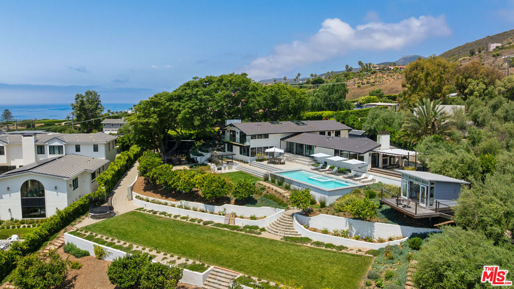 Tucked behind gates and lush privacy hedges, this stunning coastal smart home welcomes with manicured landscaping, stone pavers, a bubbling fountain, and a modern glass-and-metal front door. Inside, a skylit foyer with wood floors and a sitting area sets the tone for warmth and sophistication.Step up to the living room, where wide sliding glass doors frame sweeping ocean views. A stone fireplace, built-in speakers, and hardwood floors create a relaxed, elegant setting. The home is outfitted with a Lutron lighting system and Sonos audio, offering seamless automation throughout.The adjacent chef's kitchen is fully equipped with Thermador appliances including a six-burner range, BBQ, double ovens, and warming drawer. A spacious island with bar seating, mini fridge, and Bosch dishwasher anchors the room. Glass-and-metal cabinetry, built-ins, and a cozy breakfast nook with a skylight complete the space.Continue to the two-car garage with custom storage and the laundry room with stone counters, Samsung appliances, and sink. Nearby, a private lower-level suite offers a spacious bedroom and separate living/play area with a spacious closet, backyard access, ocean views, and a fireplace. The en suite bath features marble counters, a tiled shower with dual showerheads, and custom built-ins.Upstairs, the formal dining room opens to a tranquil patio with a fish fountain. Along the hall, a guest bedroom features hardwood floors, a hanging chair, built-in desk, and triple French-door closets. A nearby bath offers a skylight, marble floors, rainfall shower, and custom stone vanity.At the end of the hall, the primary suite is a true retreat. Vaulted, beamed ceilings crown a spacious bedroom with wood floors, a sitting area, and double-sided fireplace. Glass sliders open to a private patio with ocean views, a sectional lounge with firepit, Pilates reformer, and waterfall feature. Inside, the luxurious bath includes a freestanding tub, large steam shower with dual showerheads, marble makeup vanity, double sinks, and a water closet with bidet. Closet space is generous and fully built out.Outside, terraced stone paths wind through citrus trees, succulents, and lush lawns. The backyard includes a Tagwood rotisserie, DCS grill, and a covered dining area beside a large rectangular pool and in-ground hot tub all with panoramic ocean views. Below, another lawn leads to fruit trees and a shaded stone dining nook. Nearby is a full animal enclosure currently housing dogs, pigs, and bunnies.A detached studio office offers panoramic ocean views, wood floors, two desks, French doors, and its own deck, which is ideal for work, creative space, or a serene guest escape. Surfboard storage lies below. An outdoor shower and private sauna off the primary patio complete this extraordinary compound, where no detail is overlooked and every space is designed to elevate coastal living.
