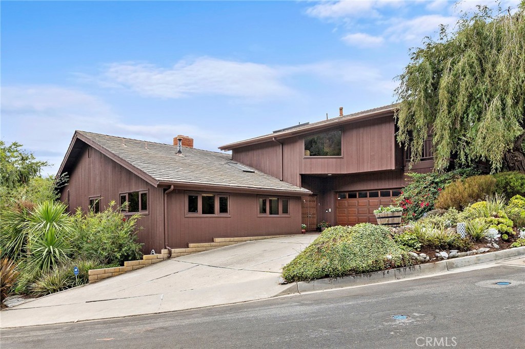 On the market for the first time in over 40 years, this Mid-Century residence embodies the timeless architecture and versatile design that are iconic to Laguna Beach. Built in 1963 and set on nearly 10,000 square feet in serene Bluebird Canyon, the home integrates wood, stone, and glass in a design that connects effortlessly to its natural surroundings. Step into the welcoming foyer, where light from vaulted ceilings and skylight above sets the tone. To the left, a private wing offers two well-sized bedrooms and a full bath. This wing also extends to a private ensuite bedroom with a walk-in closet and its own exterior entrance—ideal for a primary suite, extended family, guests, or a home office. To the right of the foyer, the dining area flows into the kitchen, where generous granite counters, ample cabinetry, and direct garage access make everyday living easy. On the lower level, the home’s grand living room makes a dramatic statement with its impressive stone fireplace, oversized windows, and French doors that open to the backyard. Adjacent to this space, a second large sitting room—with another set of French doors—offers versatility as a formal dining room, family room, or creative studio. Out back, the expansive deck invites gatherings and relaxation with multiple seating areas, a peekaboo ocean view, and a private, tree-framed yard. Upstairs, the primary suite enjoys vaulted ceilings, two private terraces, a walk-in closet, and an ensuite bath with double sinks and a striking open design that feels connected to the home’s architectural spirit. Celebrated for its original Mid-Century character, this home offers rare architectural integrity and scale. With dramatic volumes and a versatile floor plan, it provides the ideal canvas for thoughtful updates to create a truly timeless Laguna Beach retreat.